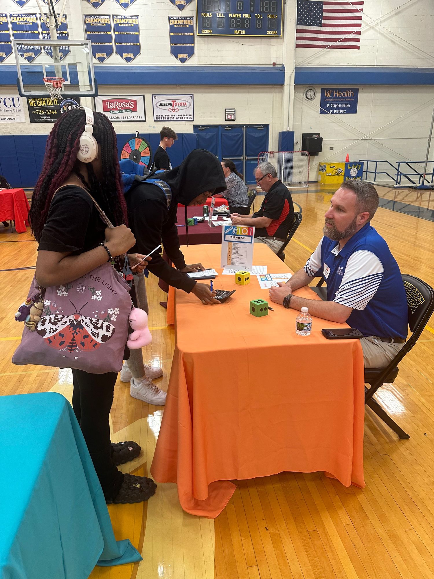 Student with headphones meets with a recruiter at an orange table during a school career fair in a gymnasium.