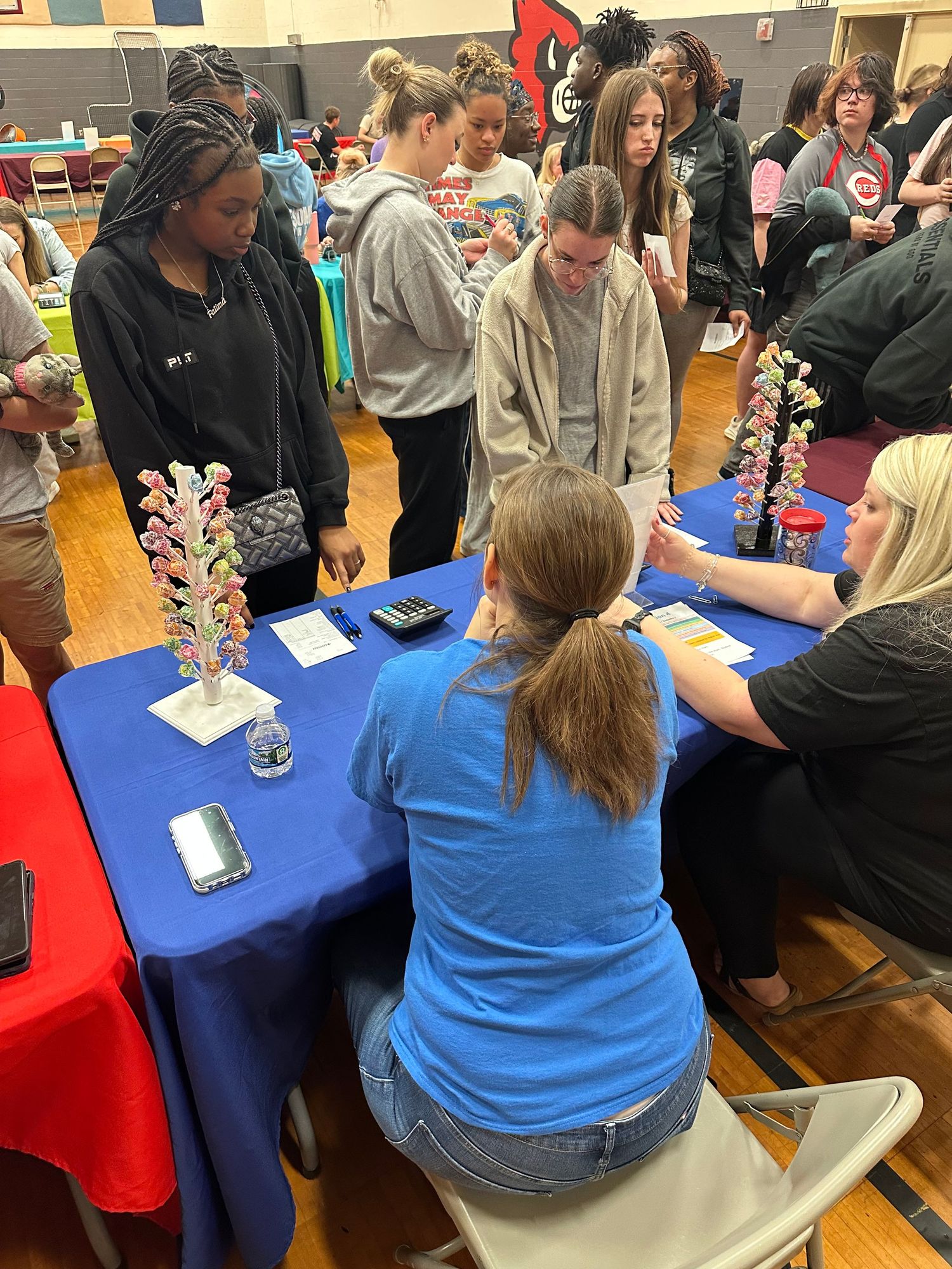 Students gather around a blue table at a school fair event in a gymnasium, engaging with displays and decorative flower arrangements.