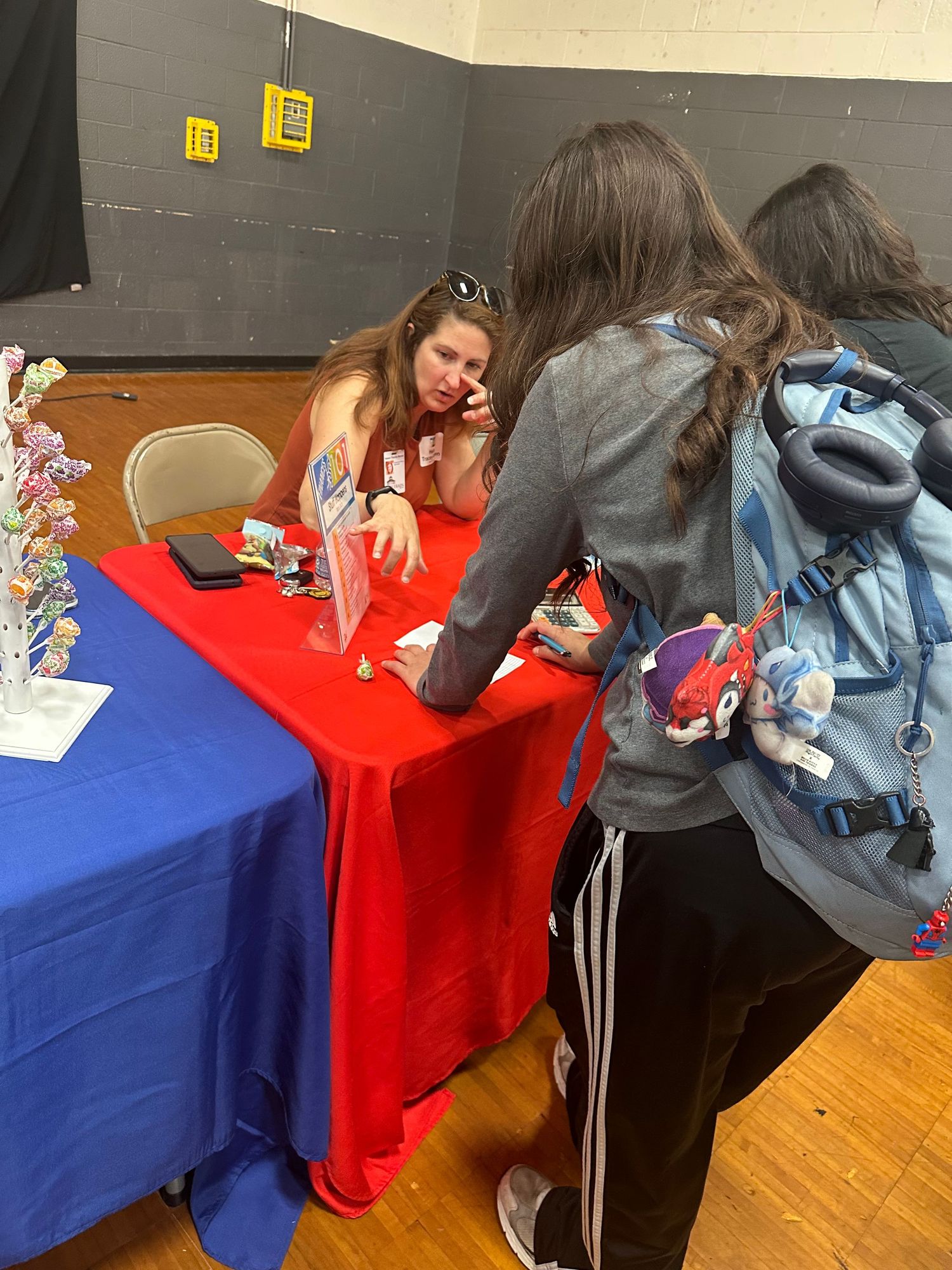 Students gather at a red and blue draped table during a school event in a gymnasium.