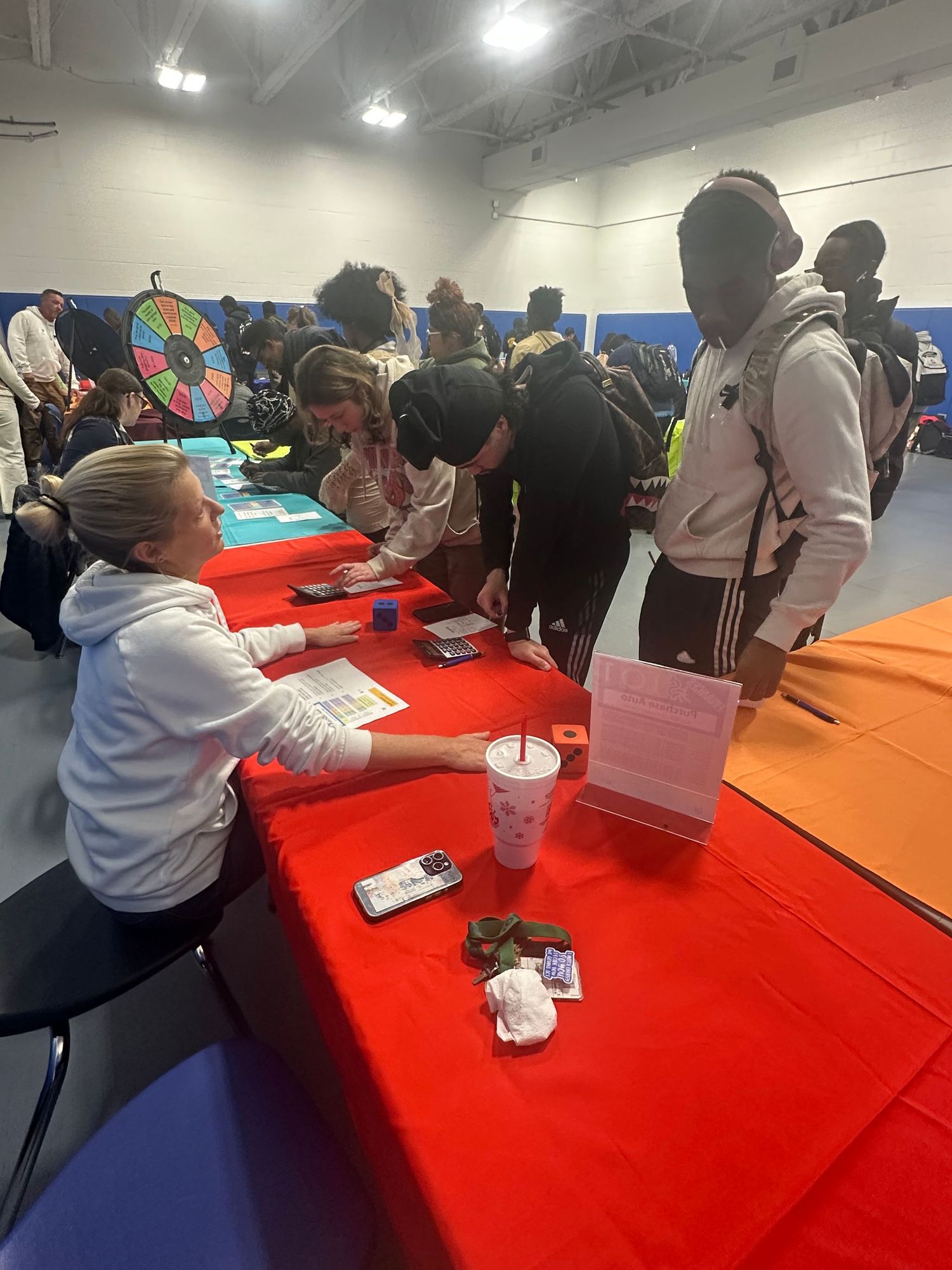 A woman assists students at a red table during a busy school career fair in a gym.