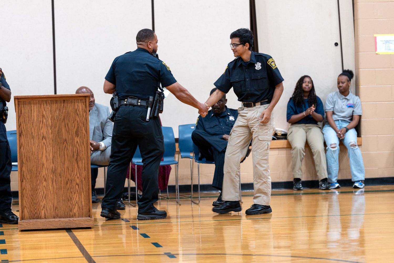 Two police officers shaking hands in a school gymnasium during a community outreach event with students seated in background.