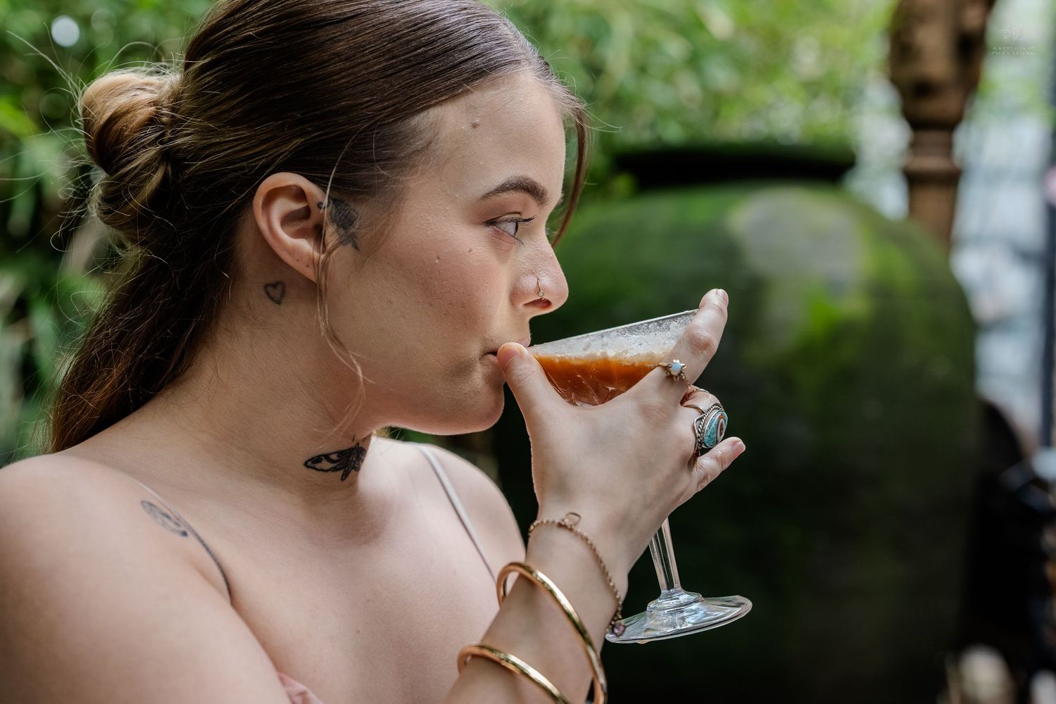Woman with hair up sipping cocktail from martini glass wearing gold bracelets outdoors.