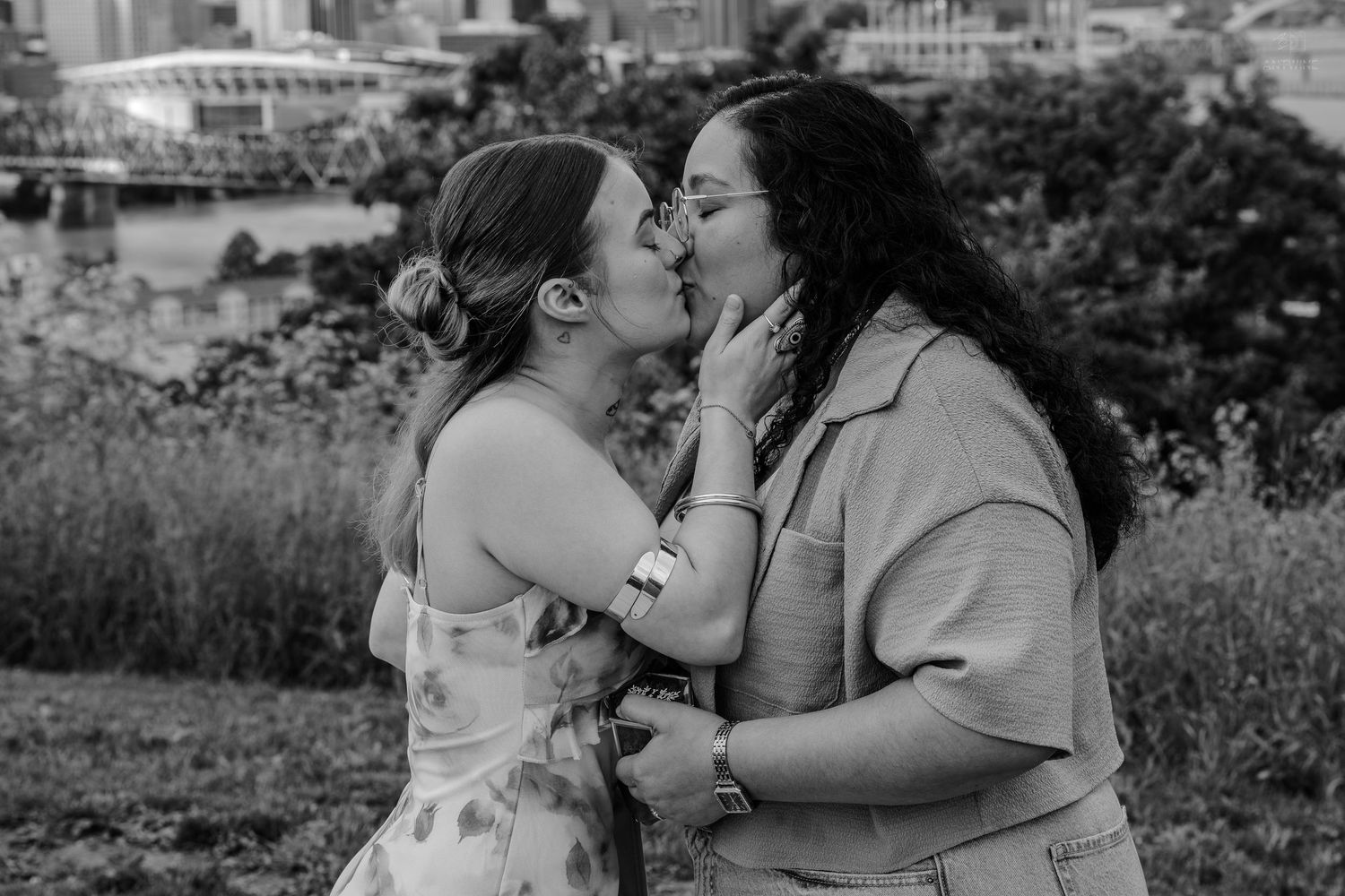 Black and white photo of two women sharing a passionate kiss outdoors with a city skyline behind them.