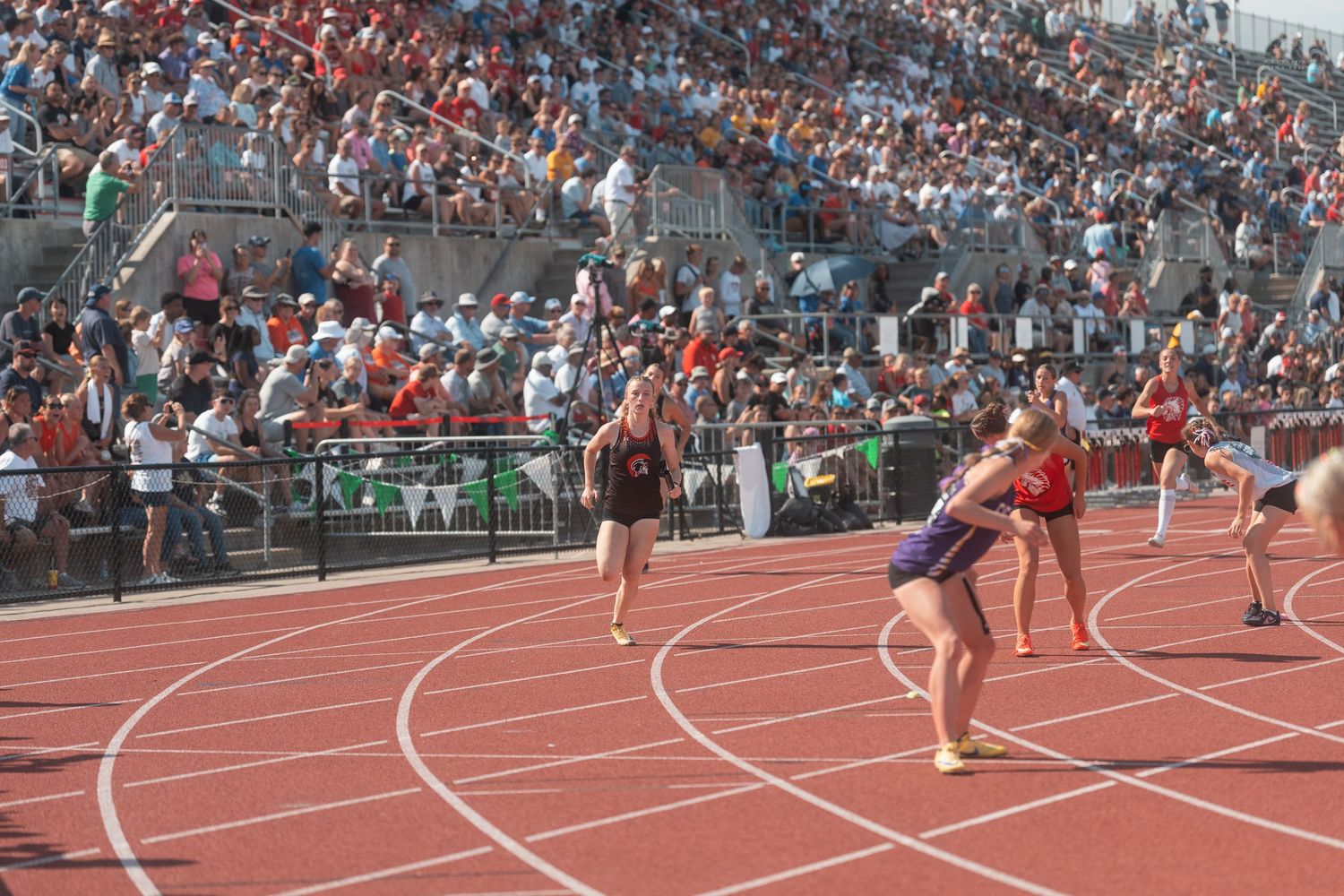 Female relay runners exchange a baton on a red track before a packed stadium of spectators.
