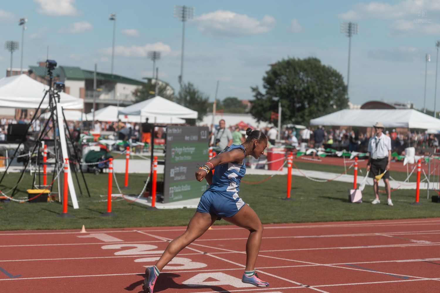 A female sprinter in blue accelerates on a red track at an outdoor high school track and field championship.