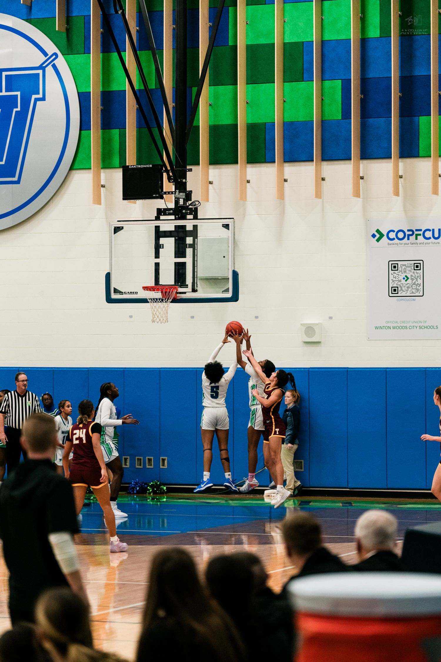 Girls basketball players jump for tip-off under the hoop during a game in a blue and green gymnasium.