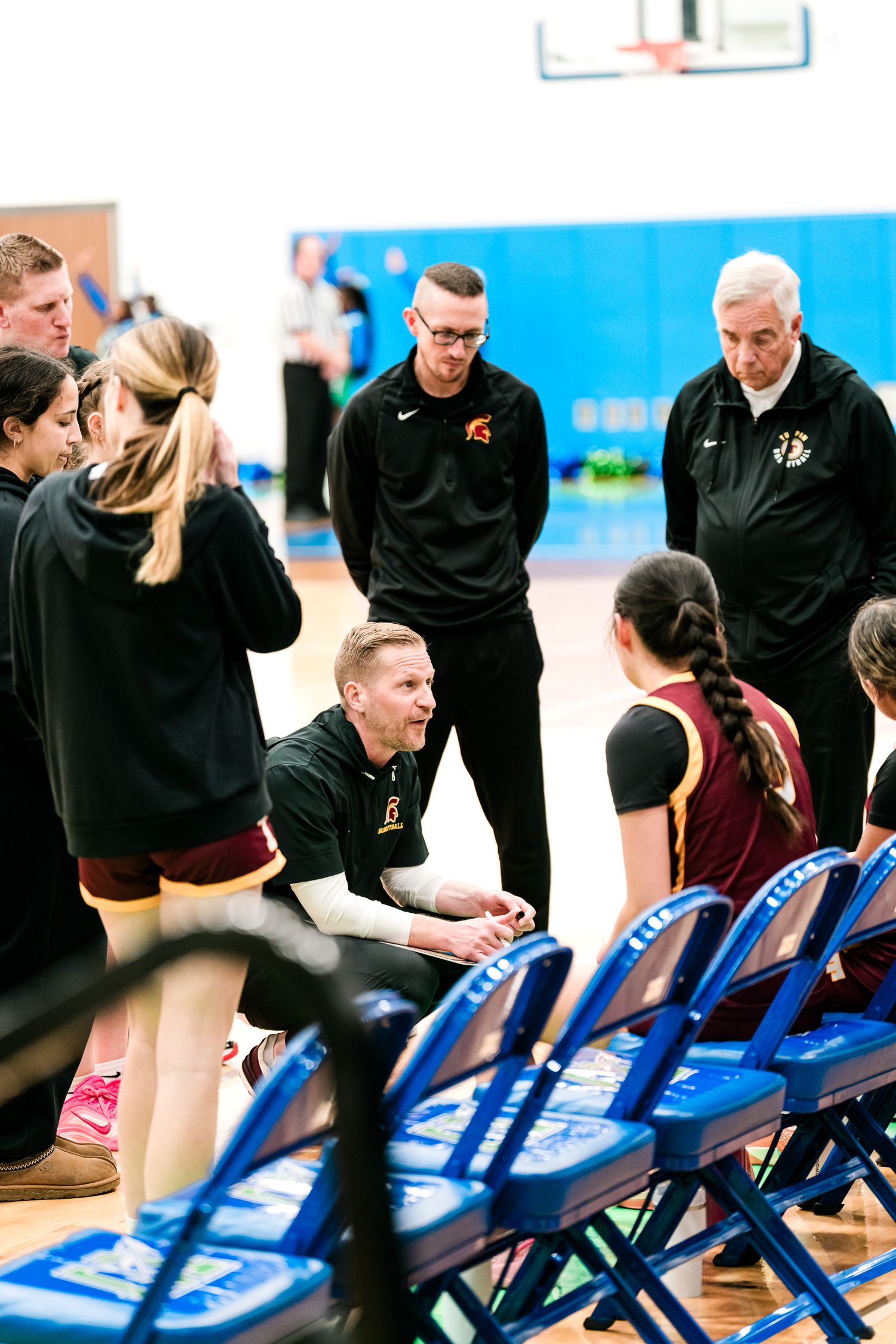 High school basketball coach crouches to address female players in maroon uniforms during a sideline timeout huddle.