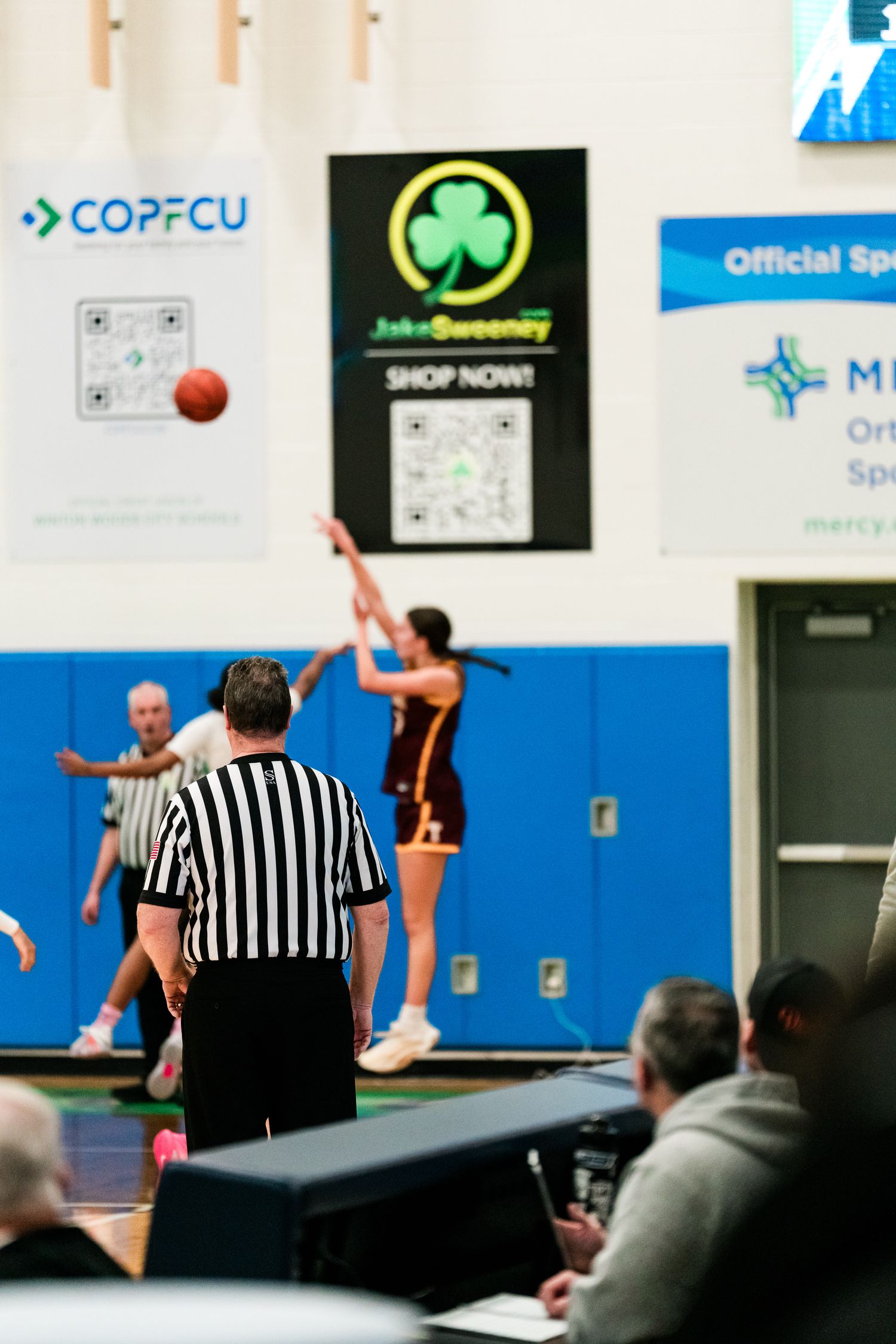 A referee watches as a player in maroon shoots the ball during a girls high school basketball game indoors.