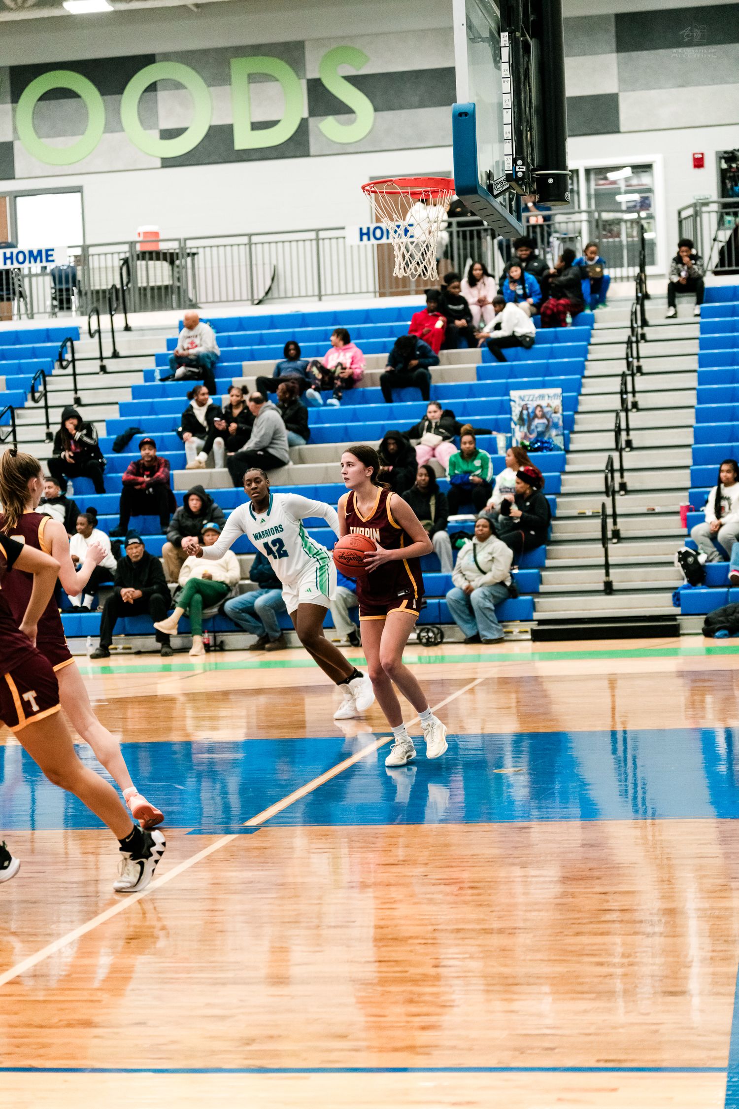 A female basketball player in maroon holds the ball while a defender in white and teal attempts to block during a gym game.