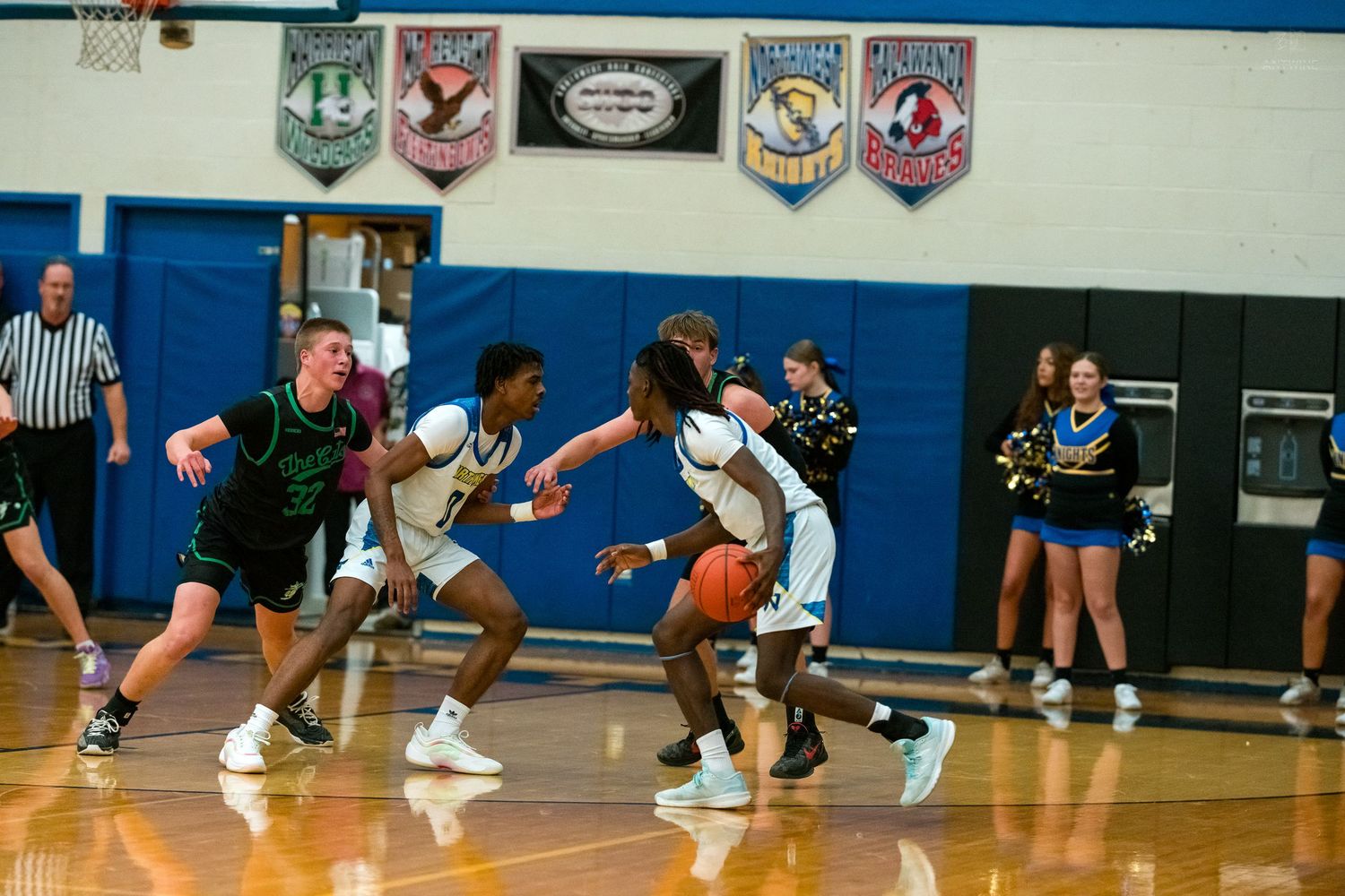 Players battle for the ball during a high school basketball game with cheerleaders and banners visible in background.