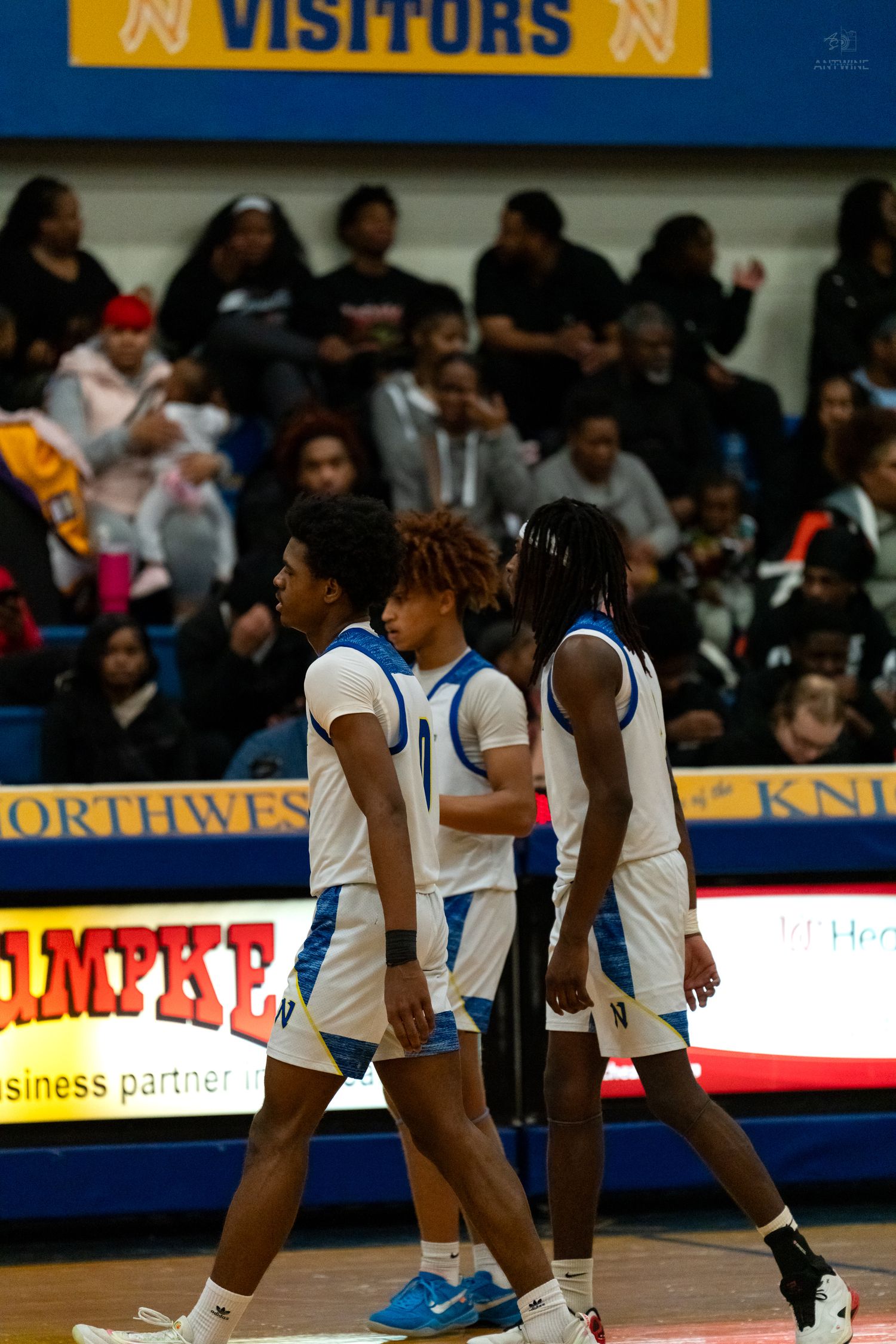 Three high school basketball players in blue and white Northwest Knights uniforms walk along sideline during game.