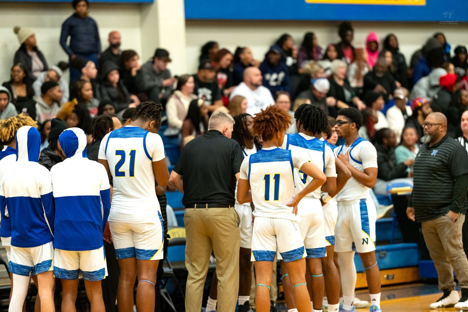 High school basketball team huddles with coach during timeout in packed gymnasium wearing blue and white uniforms.