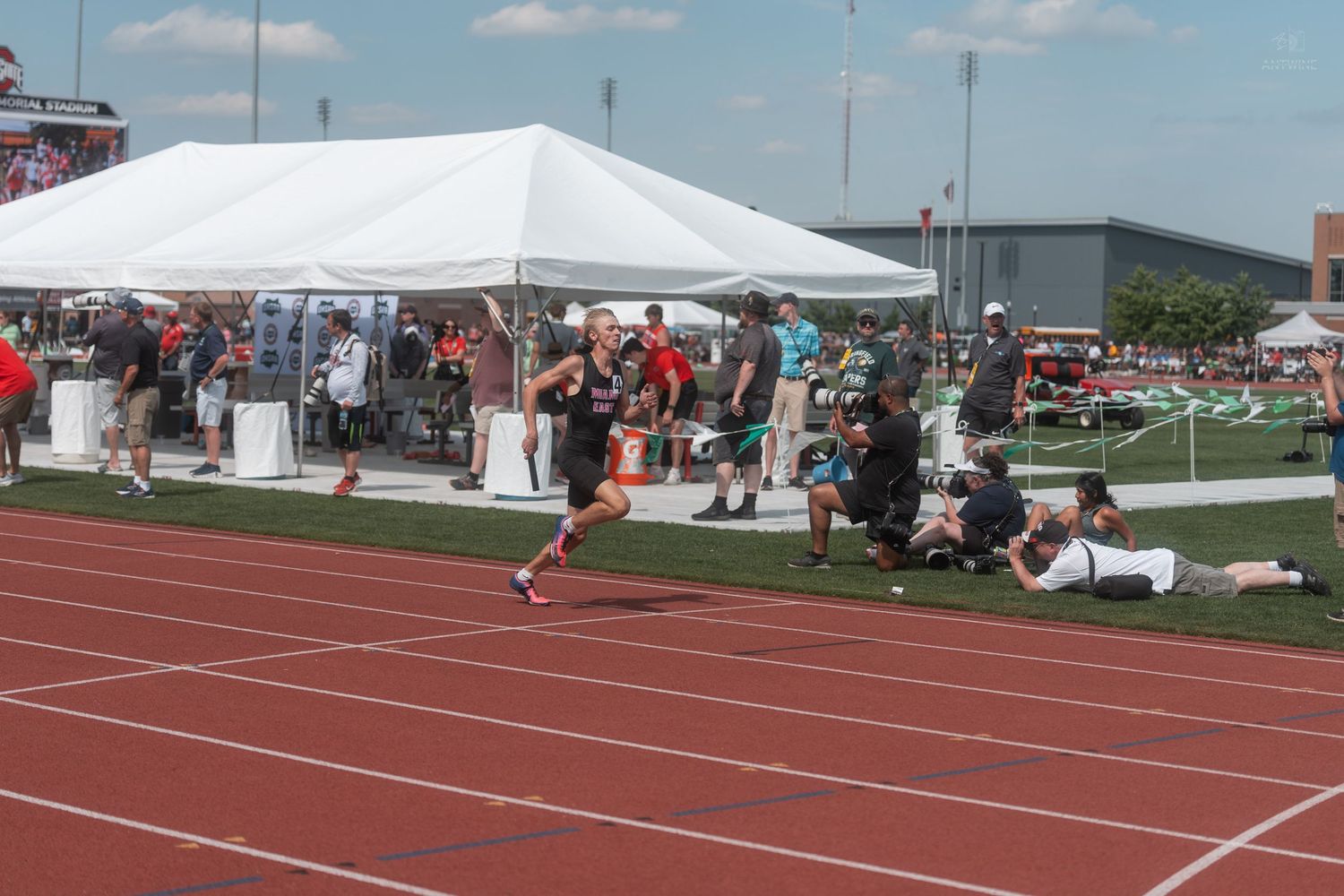 Young athlete sprints down red track at outdoor track and field event with spectators and white tents nearby.