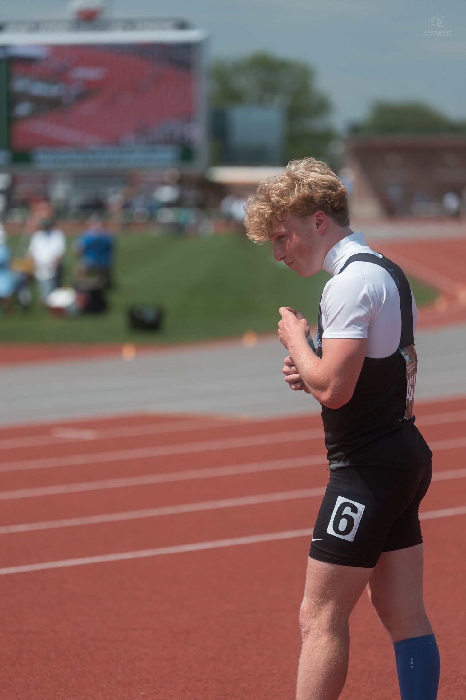 Blond curly-haired male track athlete wearing number 6 bib stands on red running track during outdoor competition.
