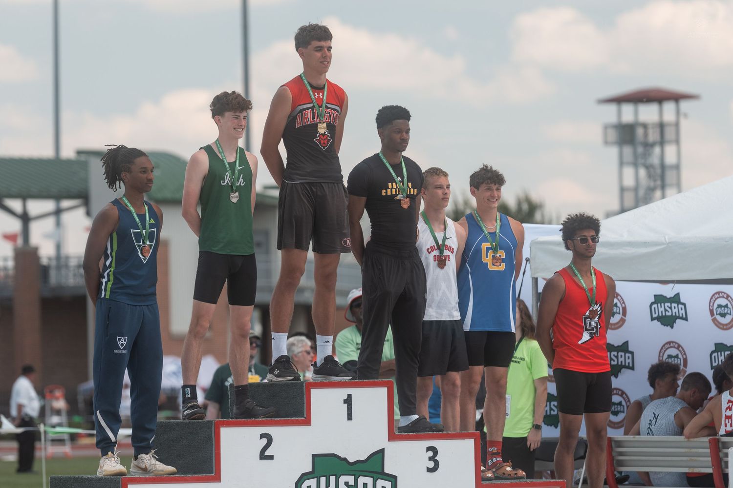 Track and field competitors stand on a awards podium displaying medals at an outdoor high school championship event.