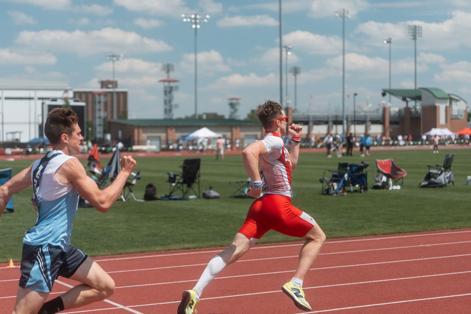 Two male athletes racing on an outdoor red track during a sunny competitive track and field event.