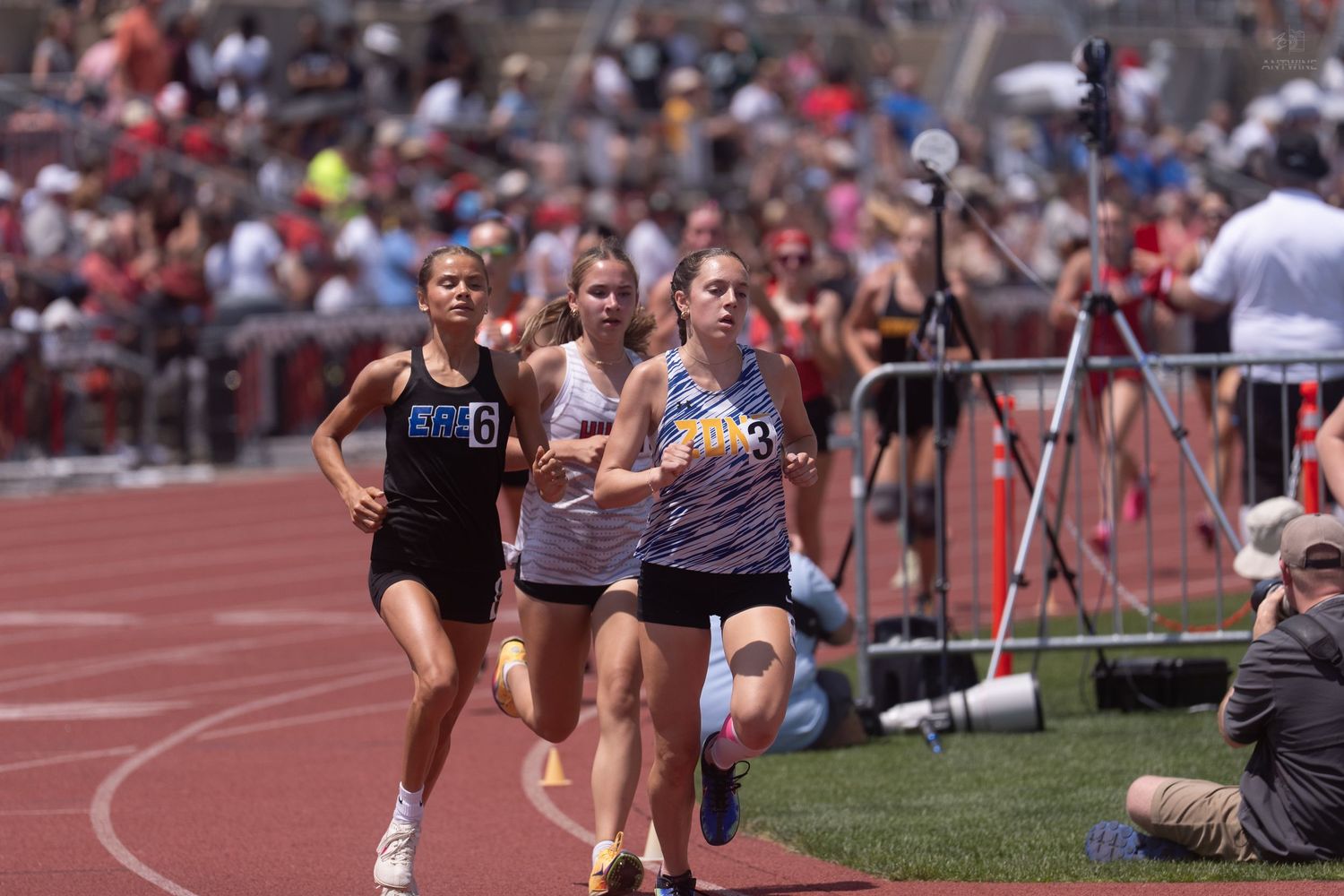 Female high school runners compete in a distance race on a red outdoor track during a sunny track meet.