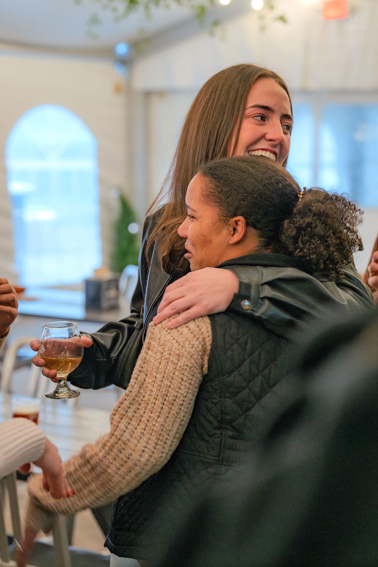 A smiling woman with long hair hugs another woman holding a glass of beer at a bright indoor social gathering.