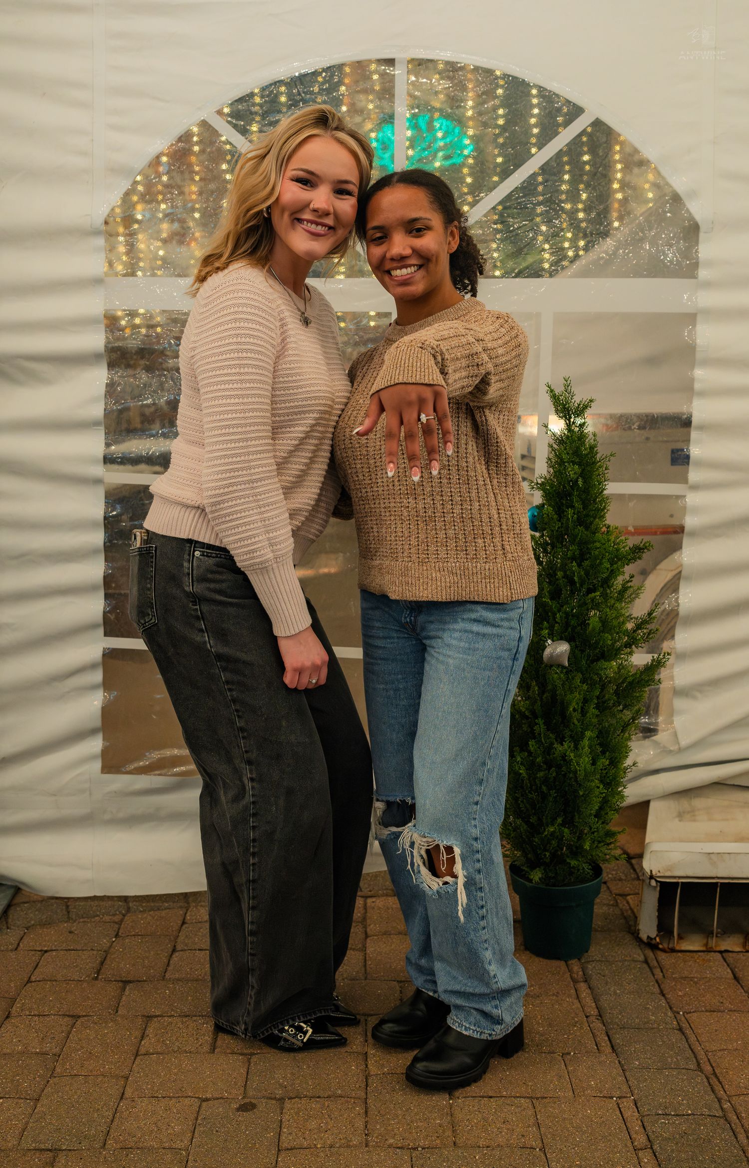 Two smiling young women posing together near a small Christmas tree inside a white event tent.