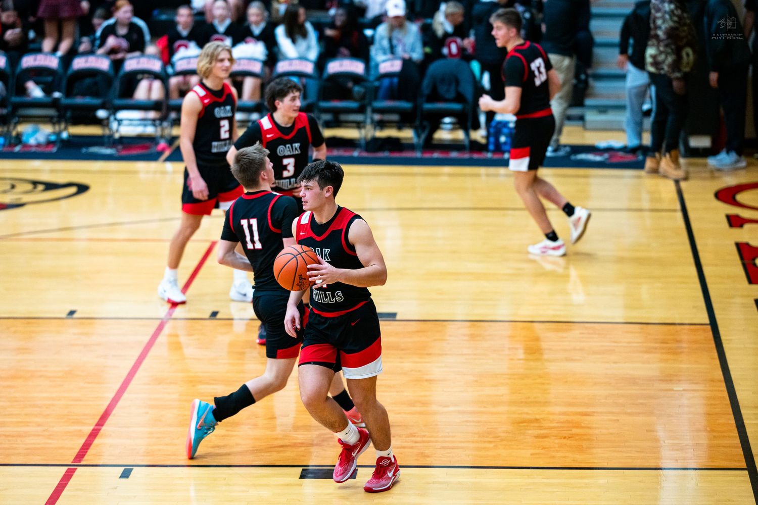 High school basketball player holds the ball while being defended during a game in a red and black gym.