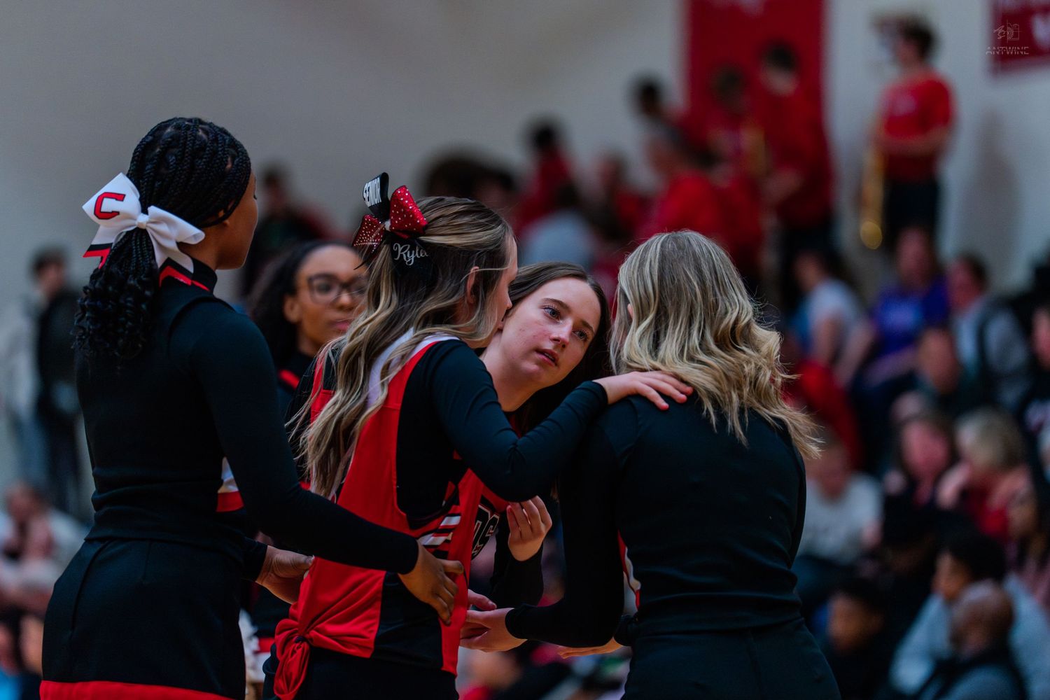 High school cheerleaders in red and black uniforms embrace during a basketball game sideline break.