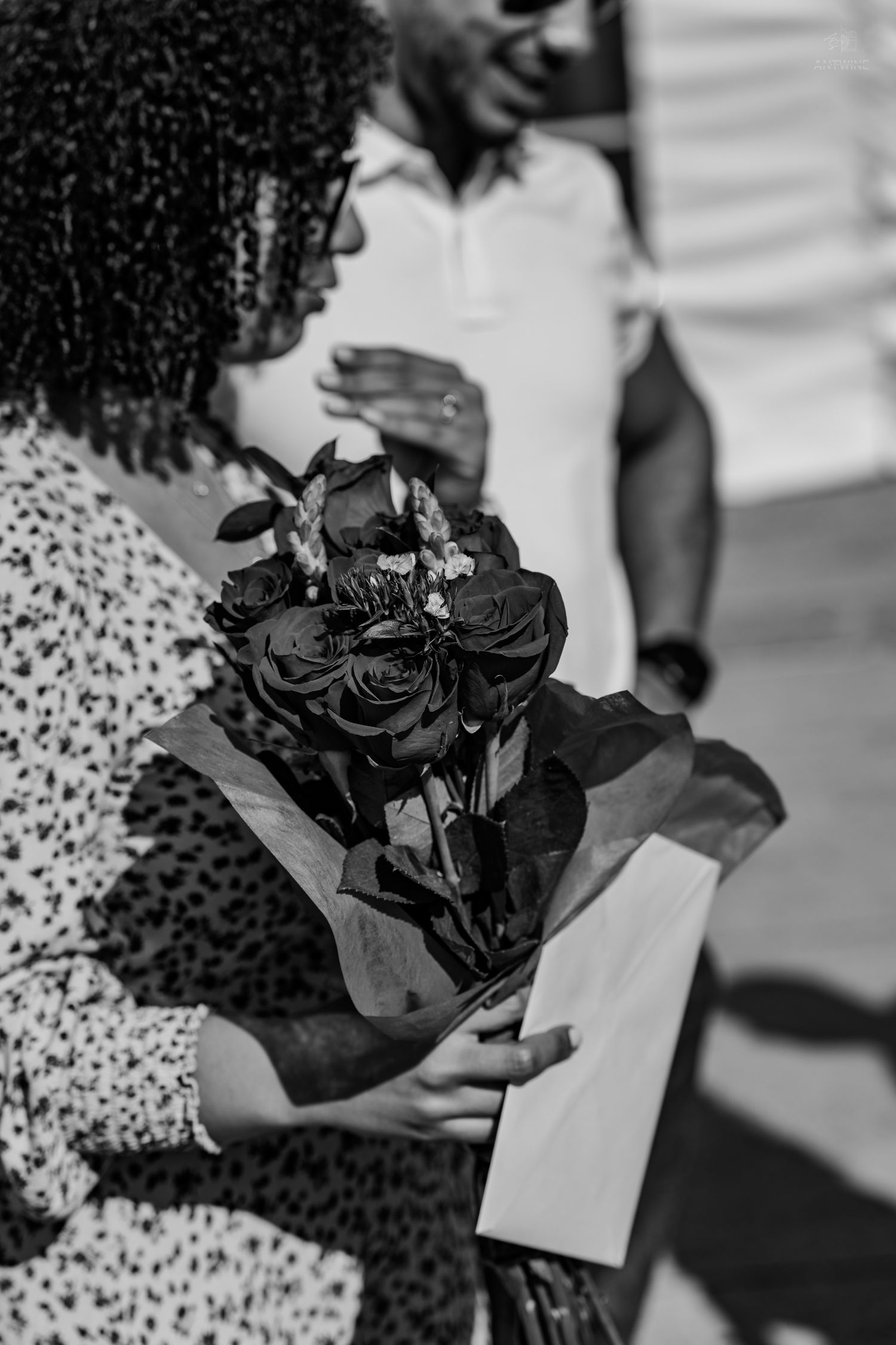 Black and white close-up of a woman holding a bouquet of roses wrapped in paper at an outdoor event.
