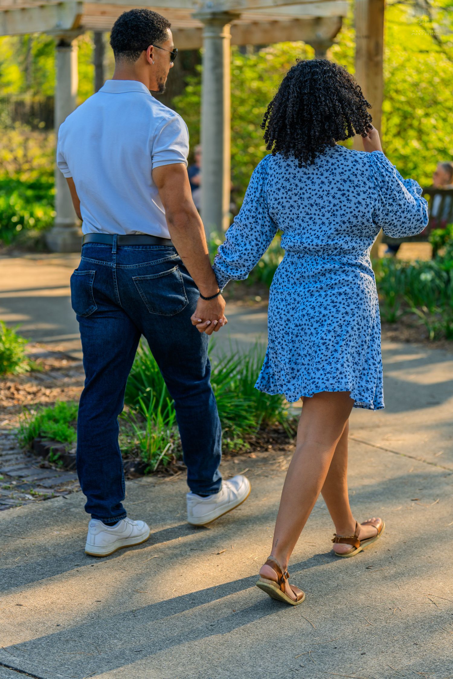 Couple holding hands walking through a lush green garden, woman in blue floral dress and man in white polo.