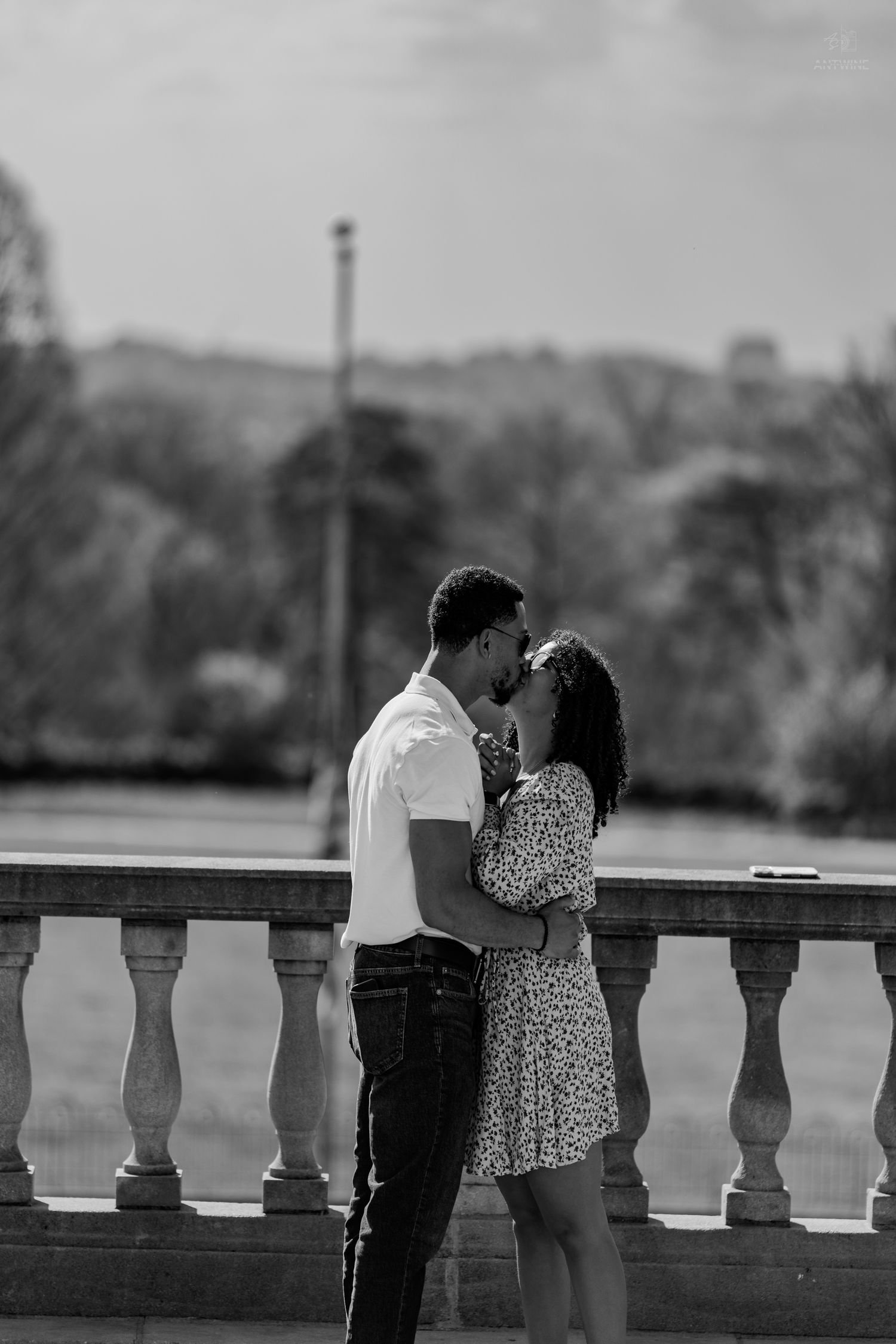 Black and white image of couple sharing intimate moment, leaning in to kiss by stone balcony railing.