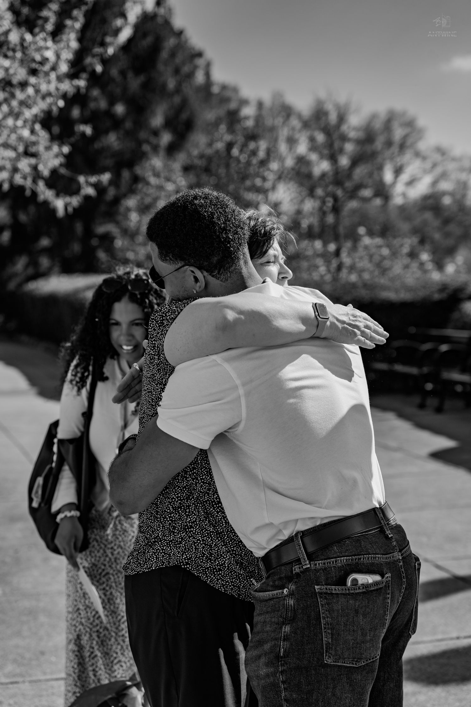Two people share a warm embrace outdoors while a smiling woman watches in this black and white photo.