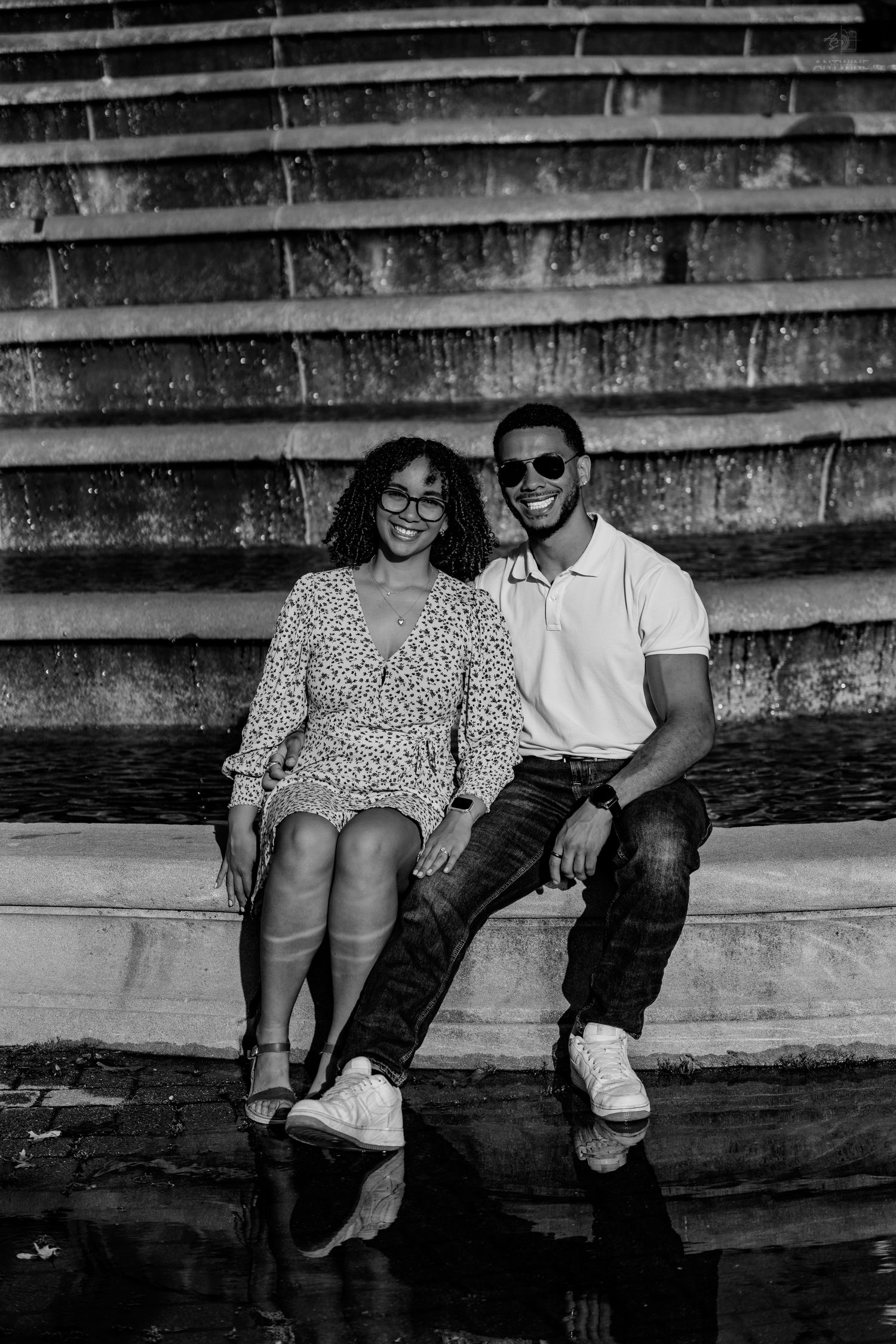 A smiling Black couple sits side by side on stone fountain steps in a black and white portrait.