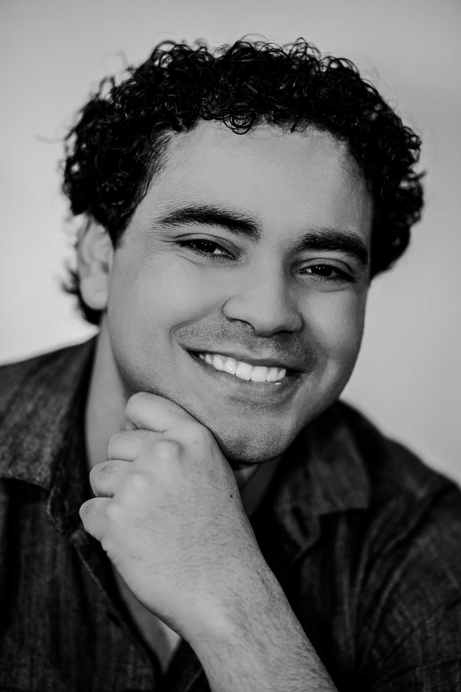 Smiling young man with curly black hair resting chin on hand in a black and white headshot photo.