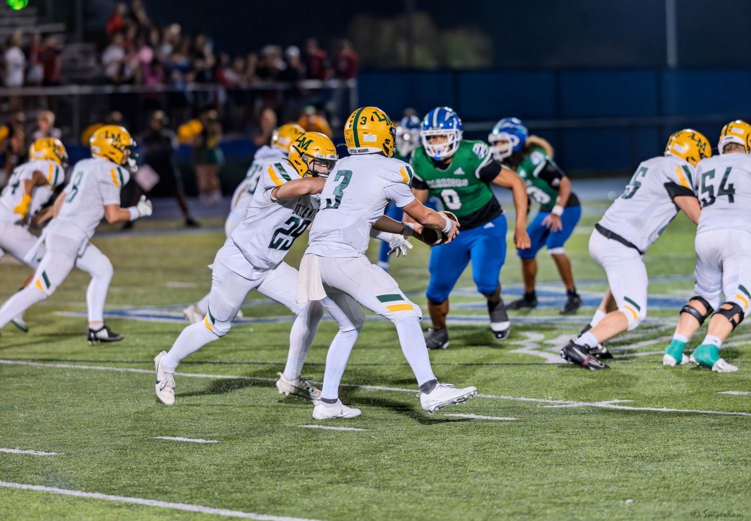 Football players in gold and white uniforms execute a handoff play while defenders in blue and green uniforms rush in.