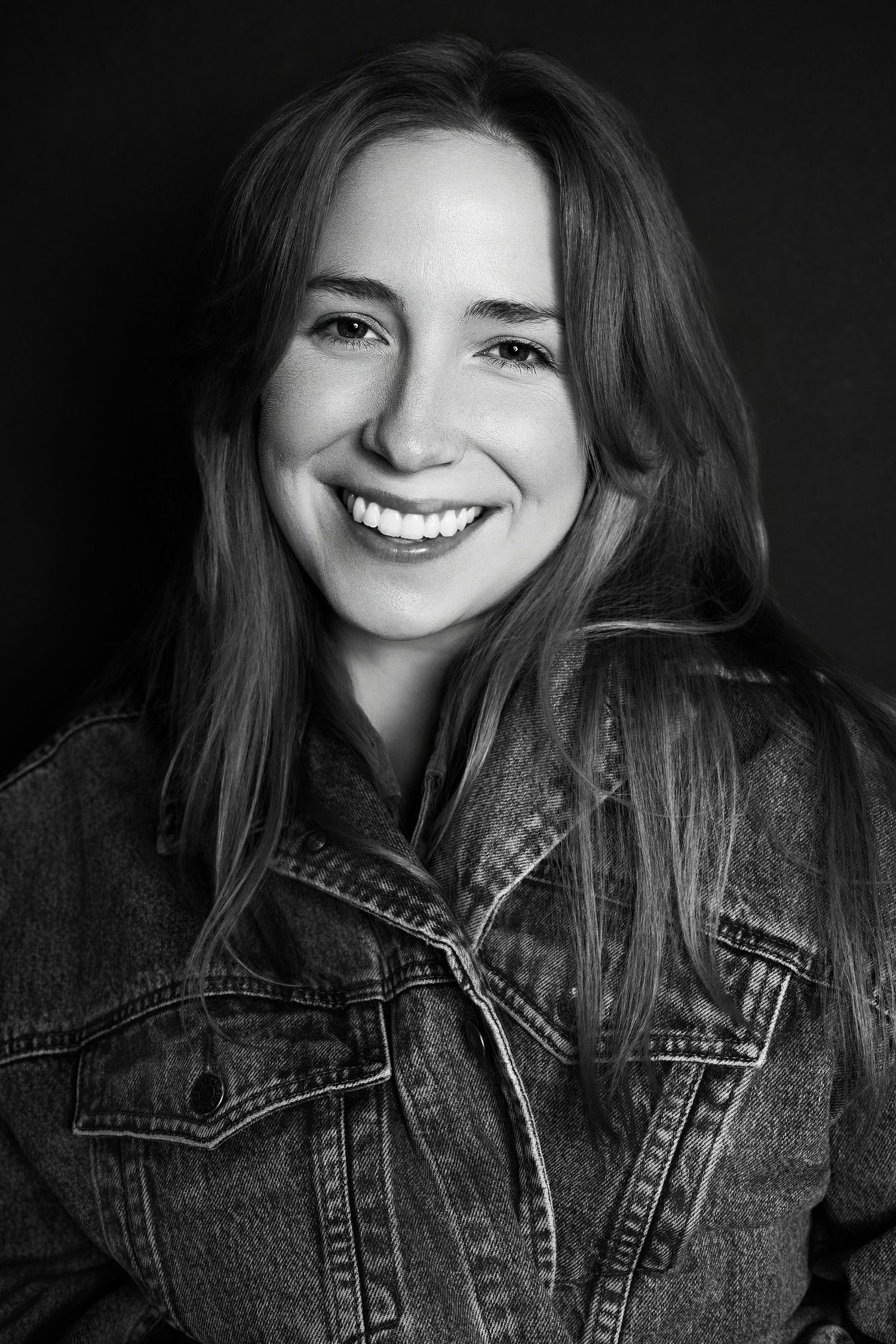 Black and white portrait of smiling young woman in denim jacket with long hair against dark background.