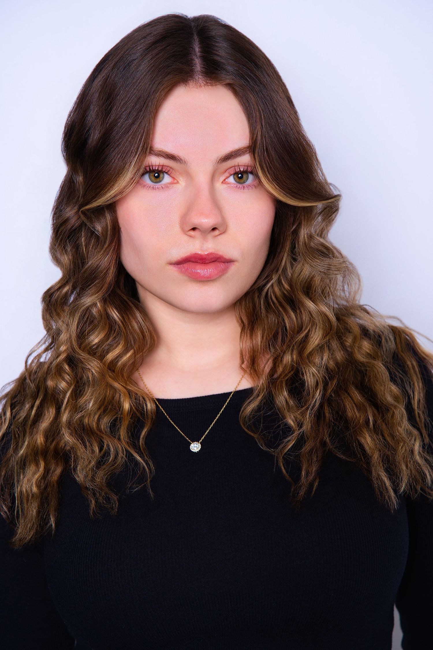 Young woman with long curly highlighted hair in black top against light gray background.