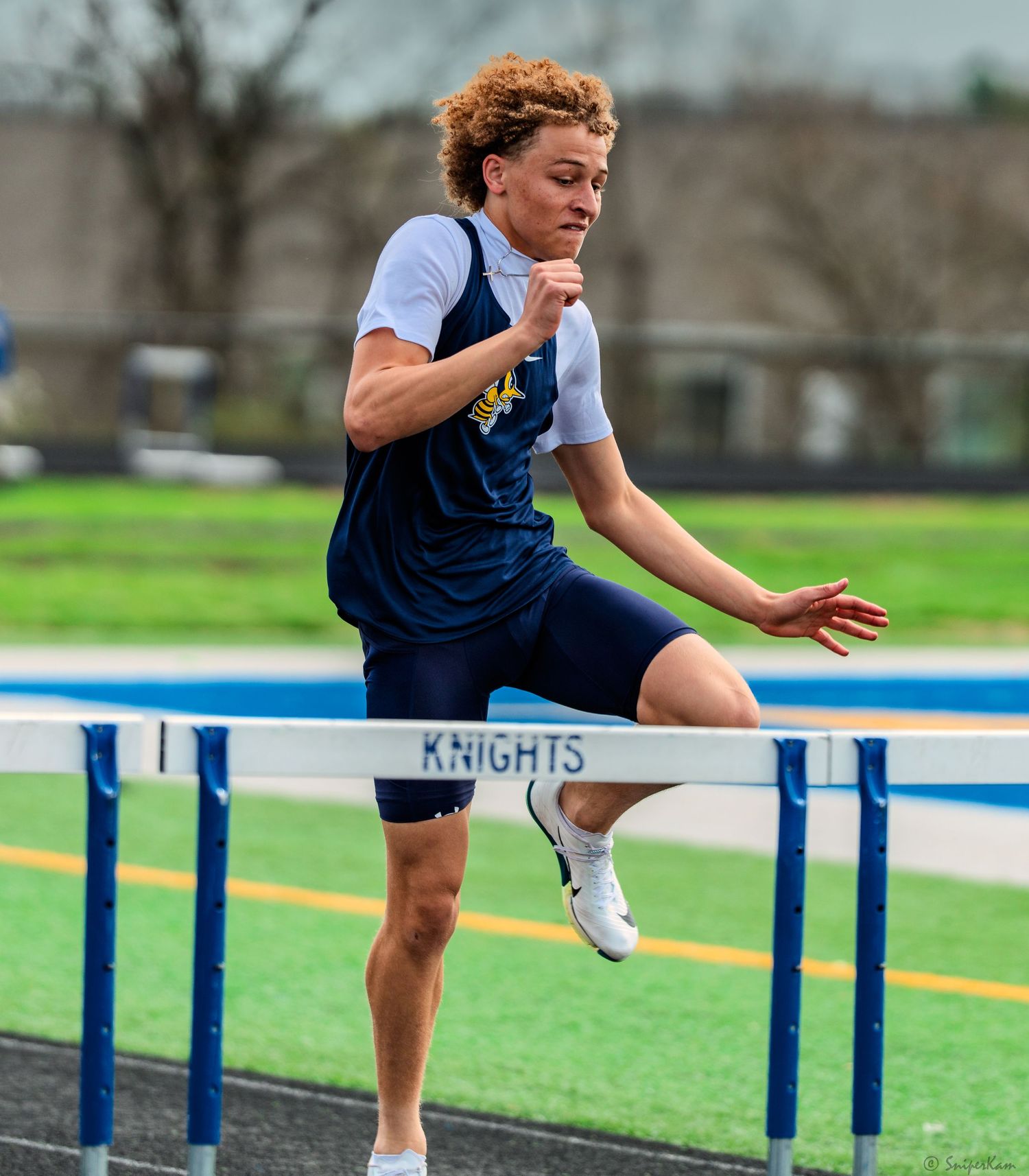 Male hurdler in navy uniform clears a Knights hurdle at outdoor track and field meet.