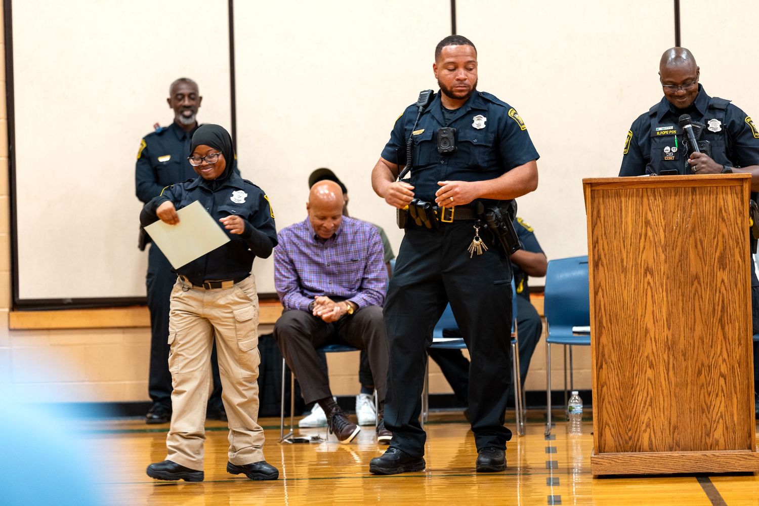Police officers stand at a podium during a community event in a gymnasium, with attendees seated nearby.