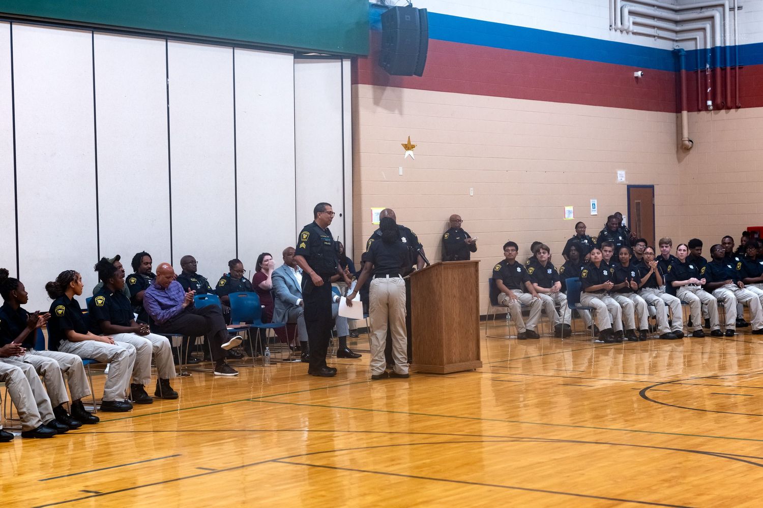 School assembly in a gymnasium with uniformed students seated along walls and speakers at a podium.
