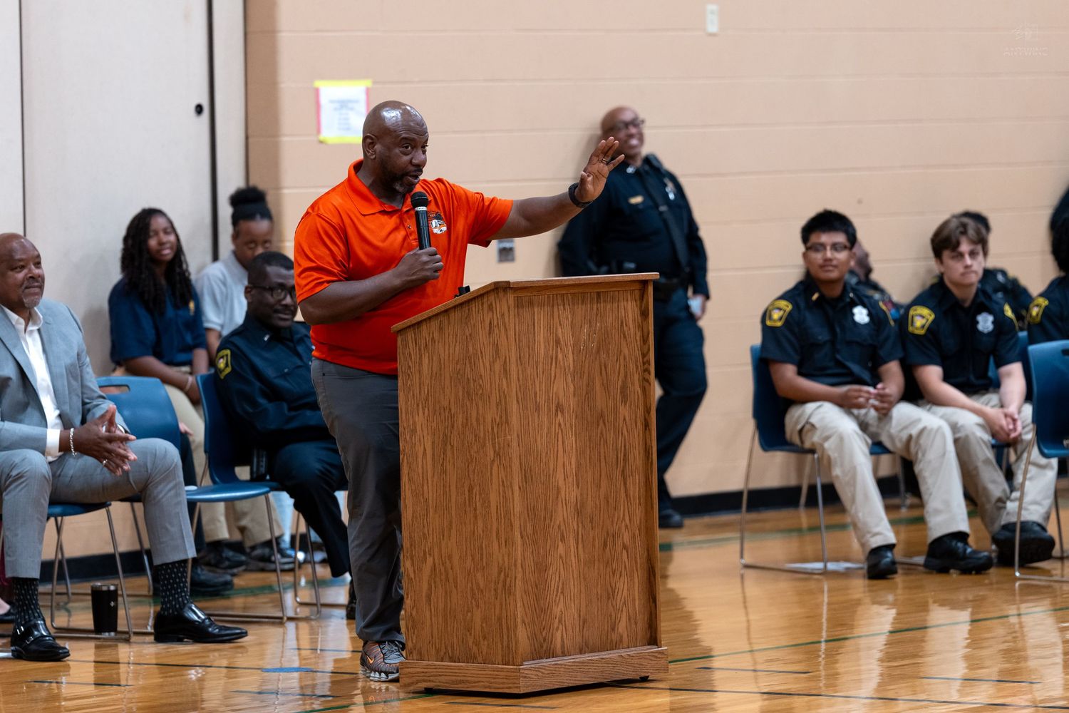 Man in orange shirt speaks at podium during community event with police officers and attendees seated in gymnasium.
