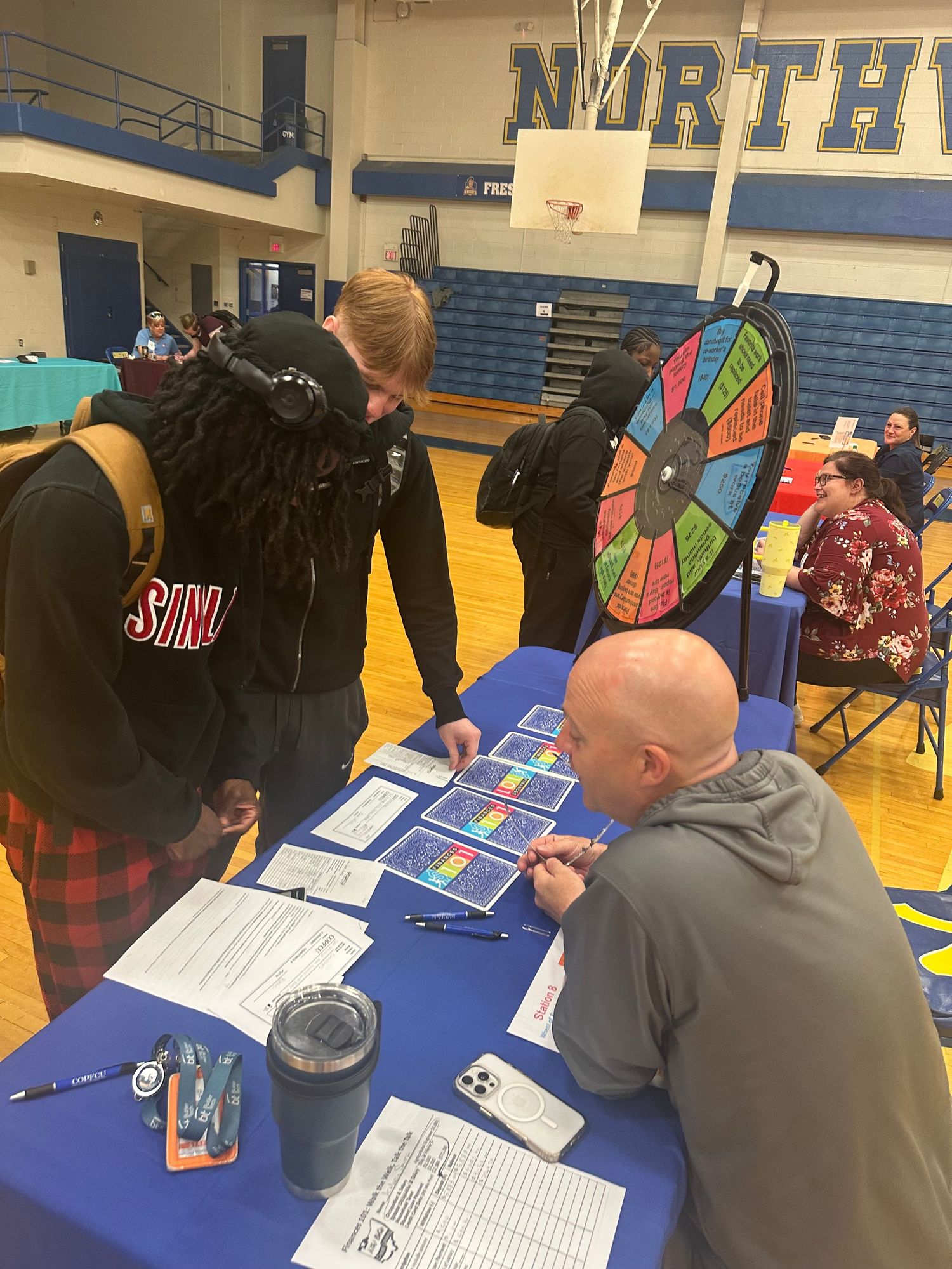 Students interact with a representative at a school fair booth featuring a colorful prize wheel in a gym.