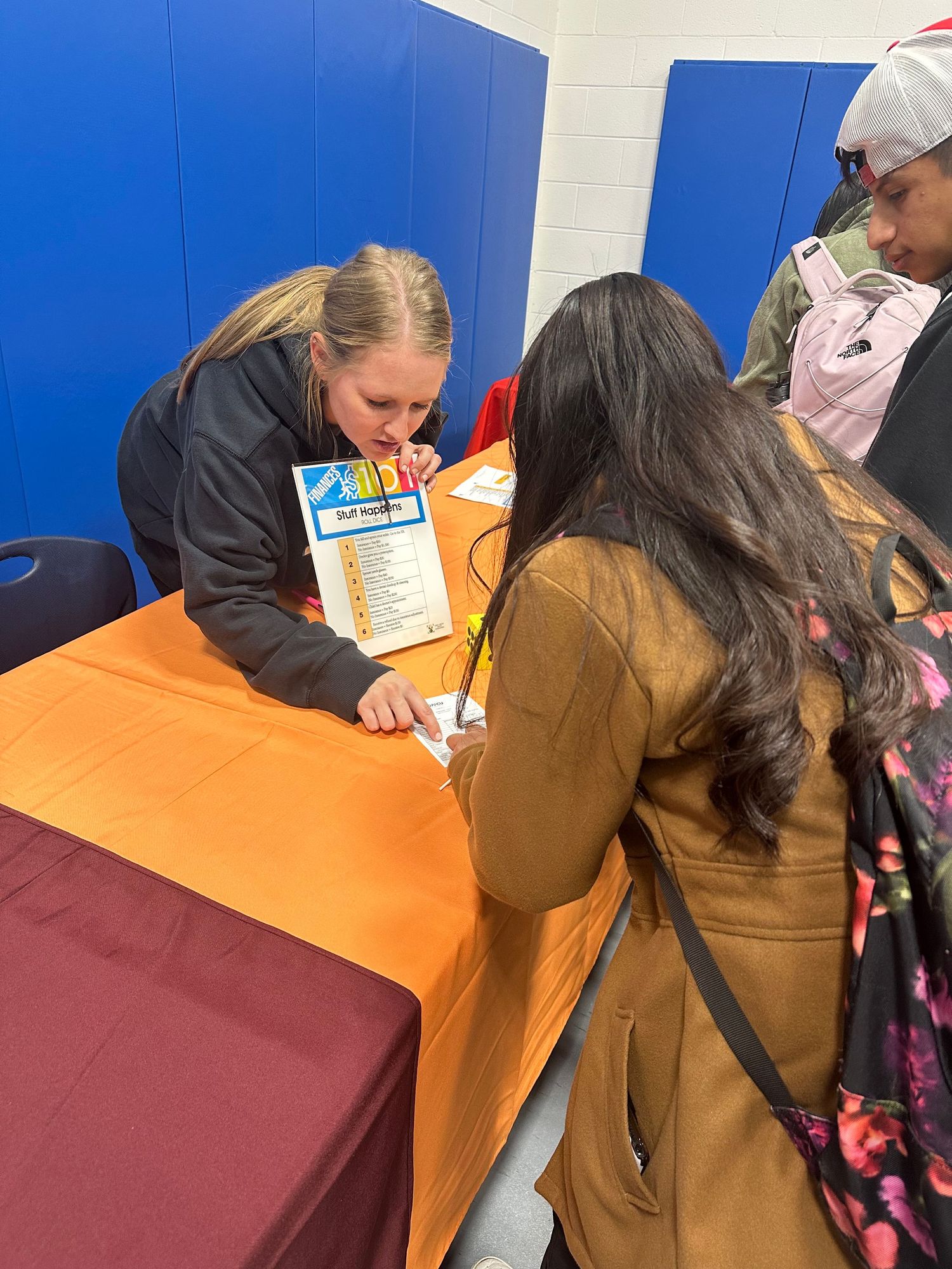 Students gather around a table at a school fair event, reviewing informational materials with a young female presenter.