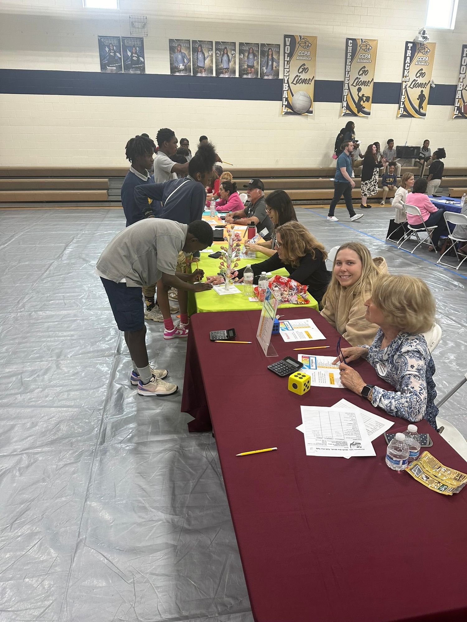 Students and adults gather at tables in a school gymnasium for an educational fair event with colorful materials.