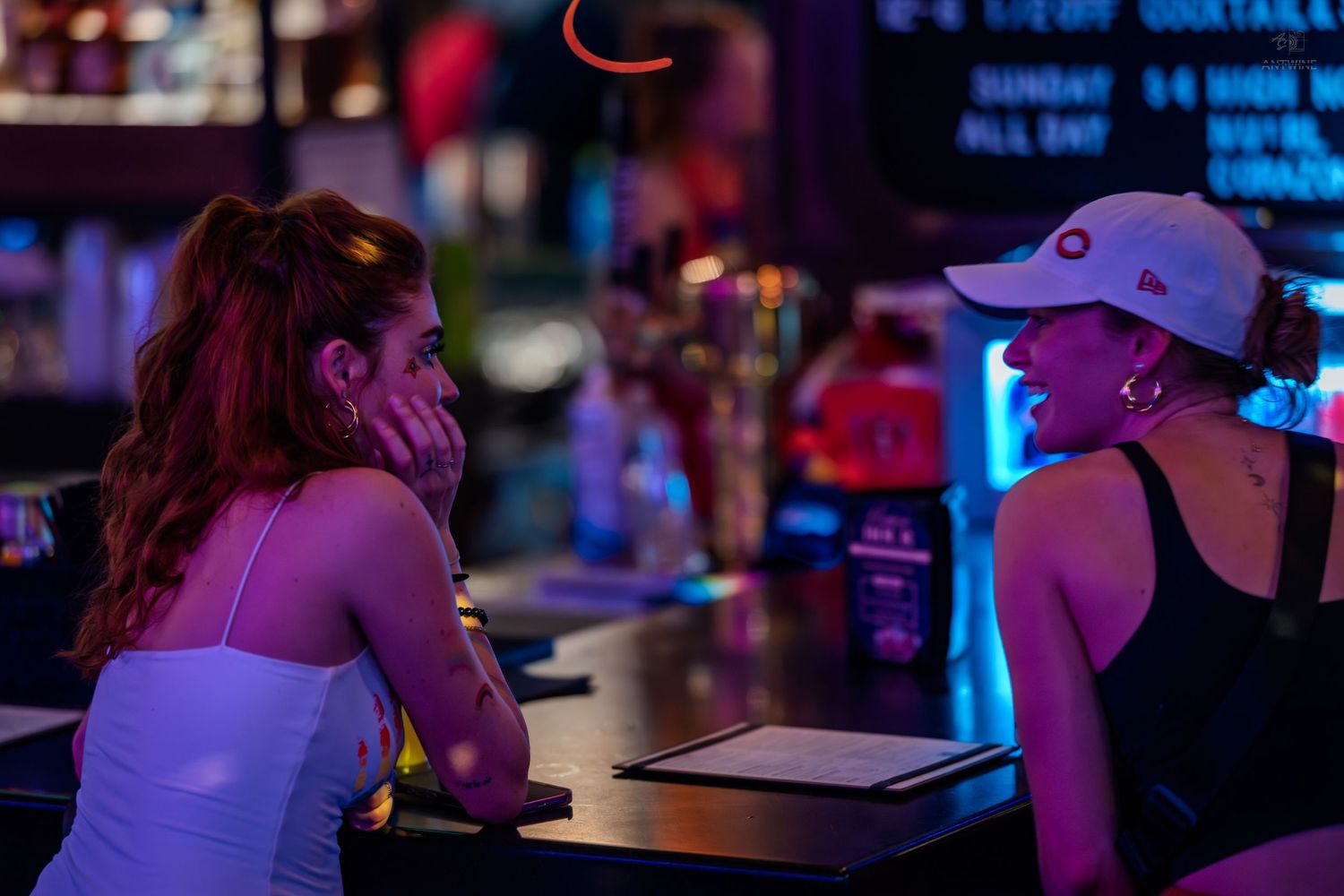Two women having an intimate conversation at a neon-lit bar counter at night.
