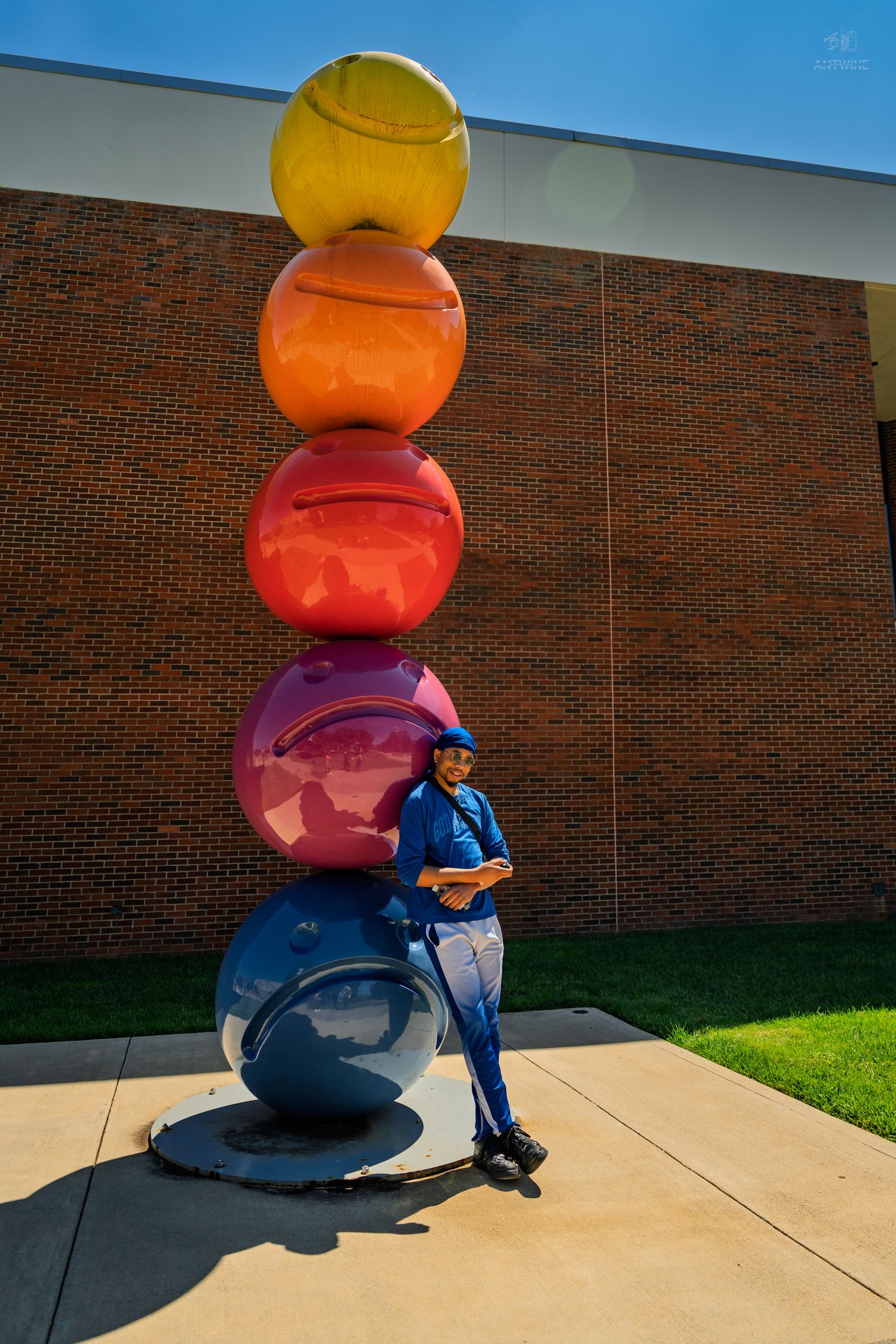 Man in blue outfit leaning against colorful stacked spheres sculpture outside brick building.