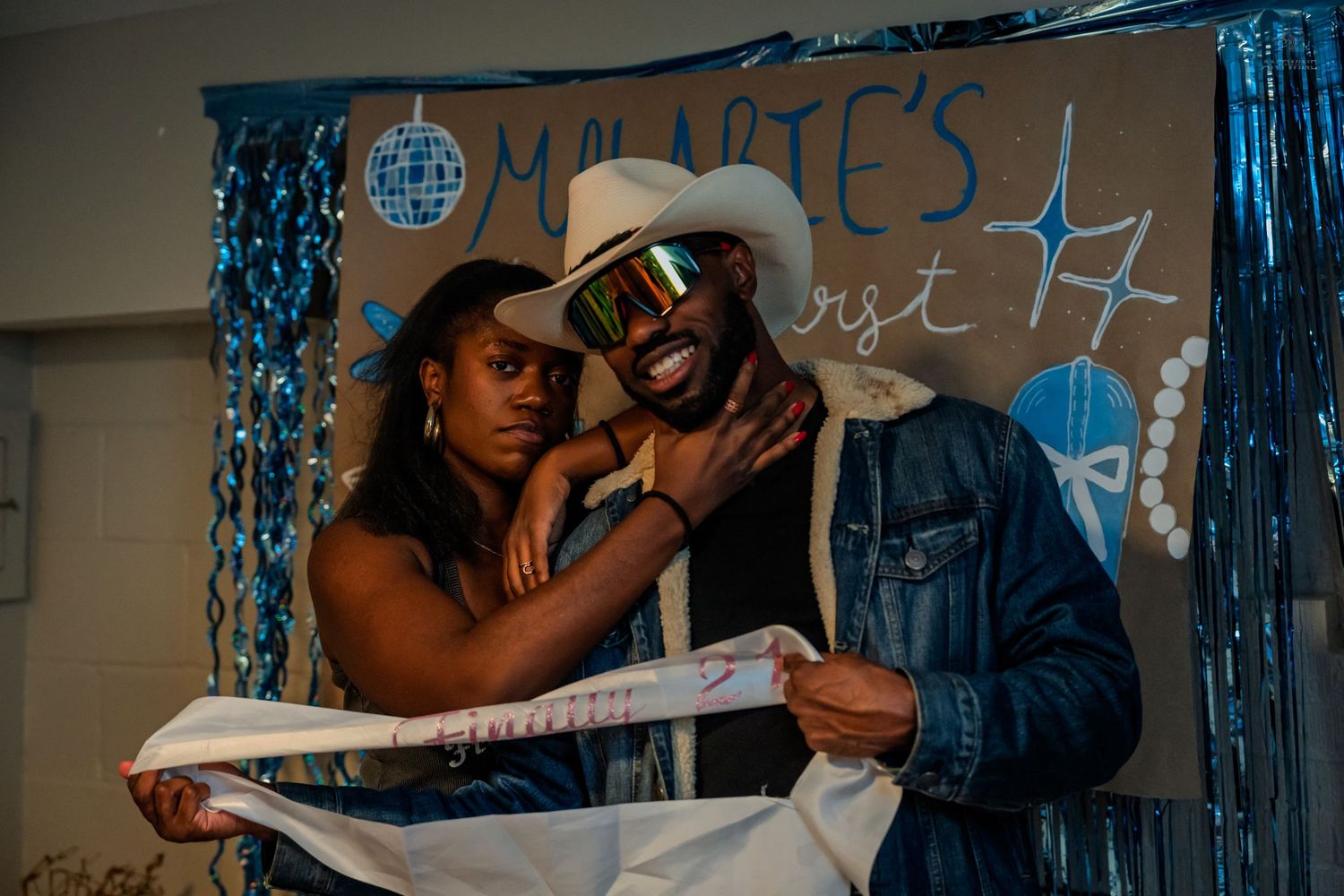 Couple smiling and holding a birthday sash together at a blue-decorated 21st birthday party.