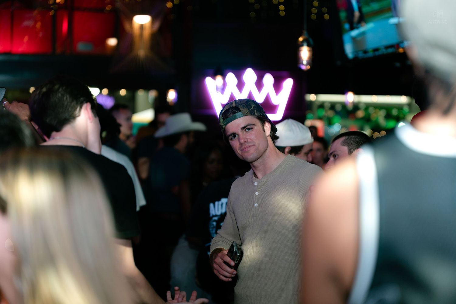 Man wearing a neon crown headband smiles at a crowded nightclub party event.