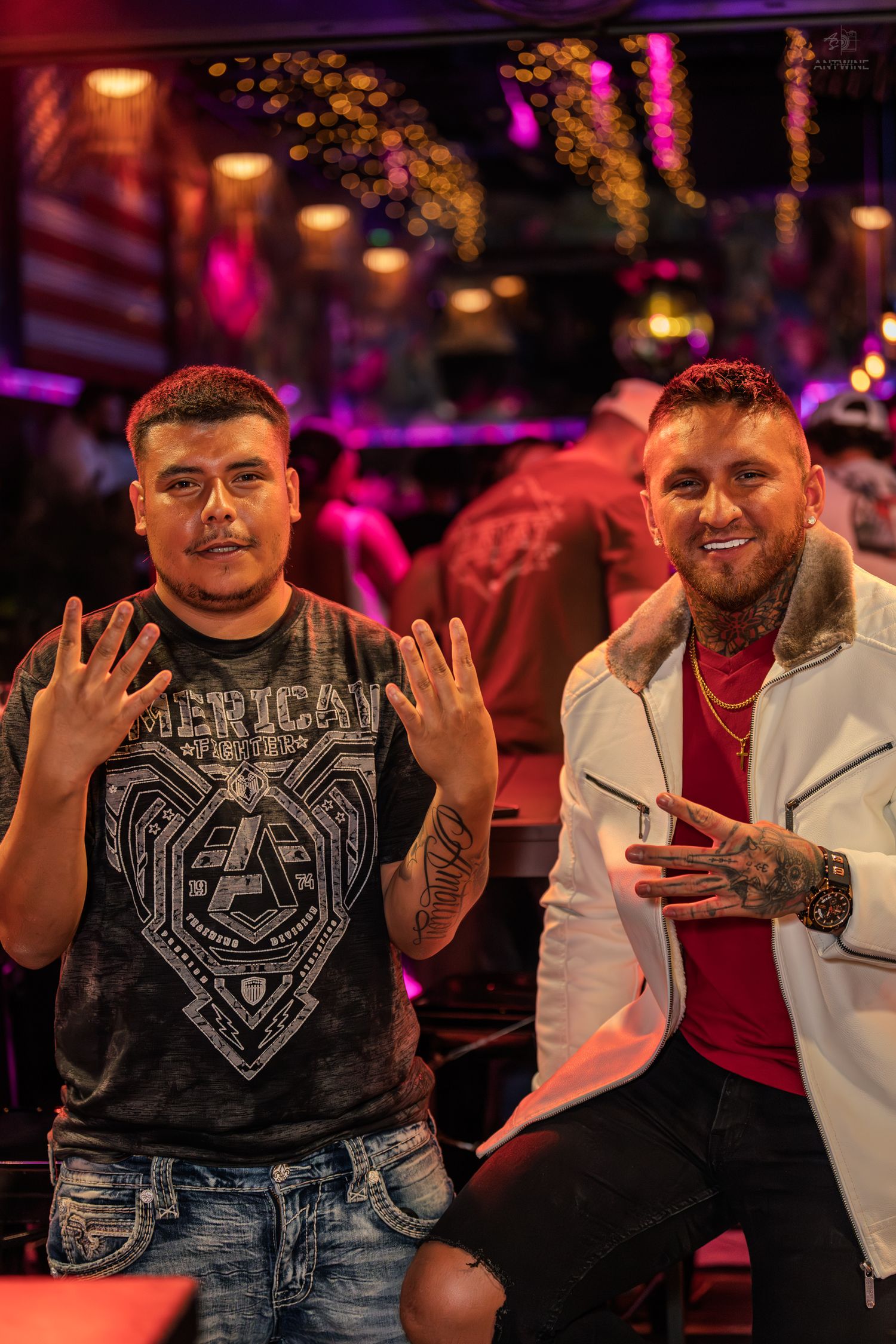 Two tattooed men smiling and making hand gestures while seated at a lively nightclub.