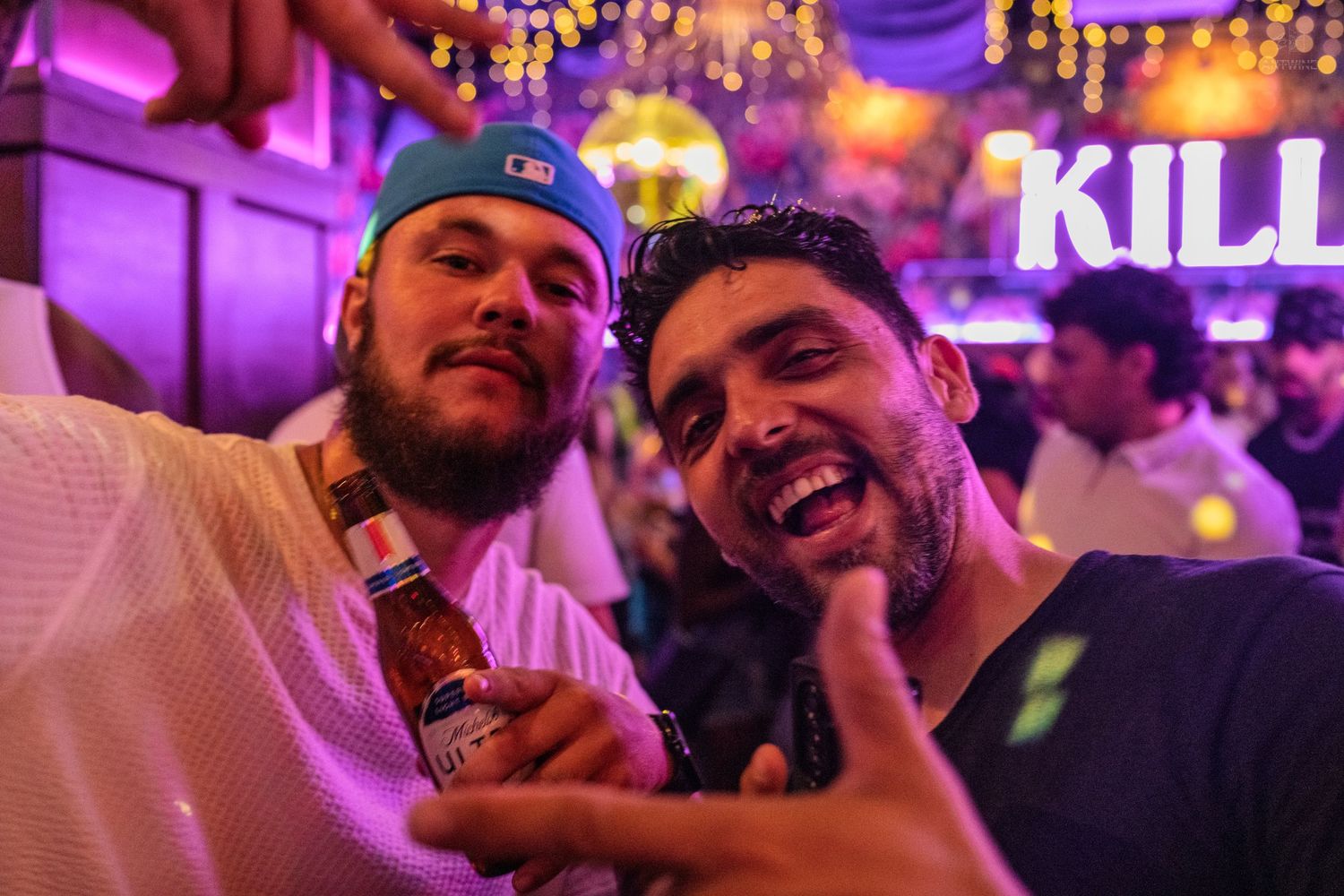 Two men laughing together at a neon-lit nightclub, one holding a beer bottle.