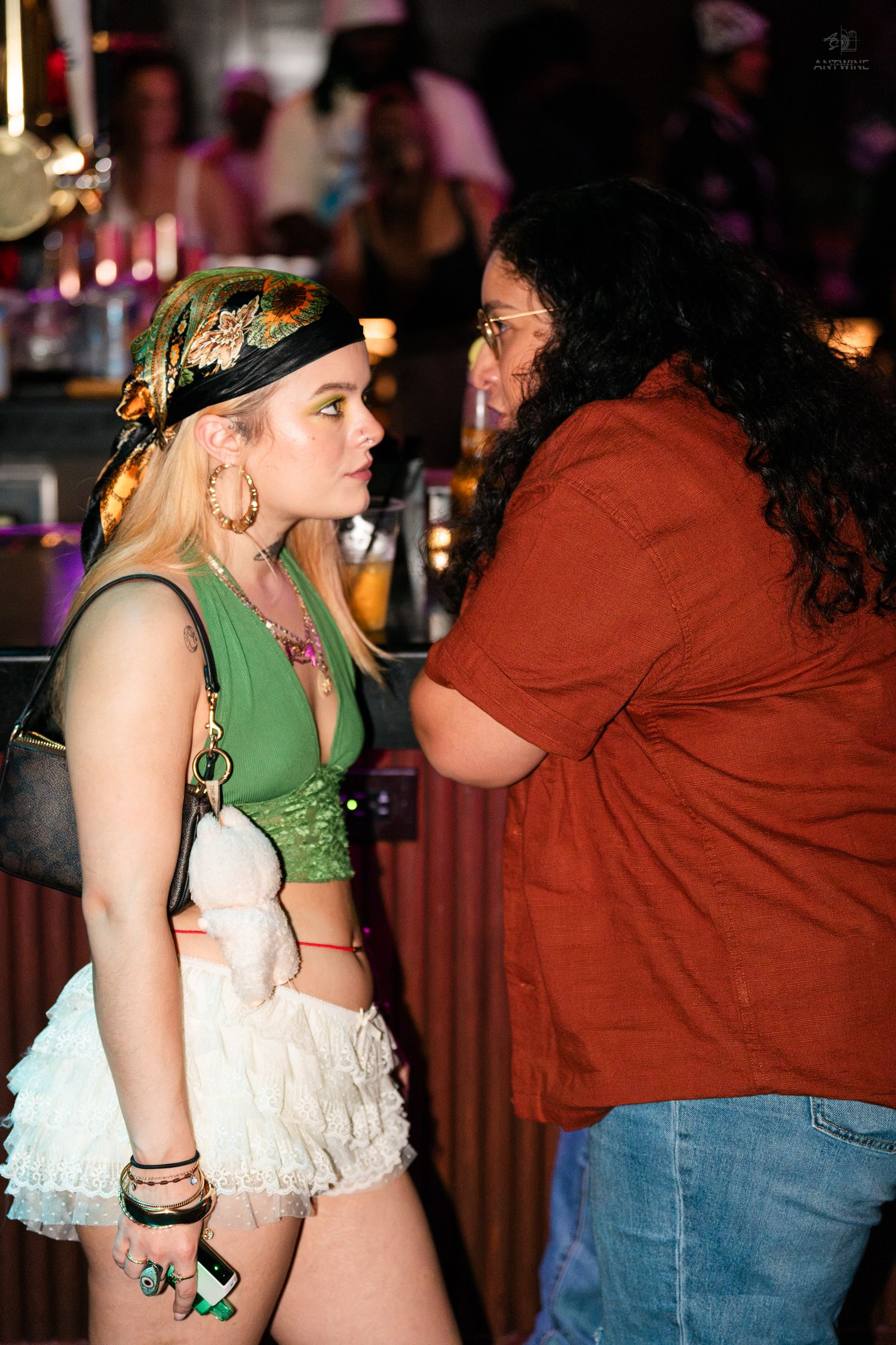 A blonde woman in green crop top and white skirt faces a woman in red at a lively nightclub.