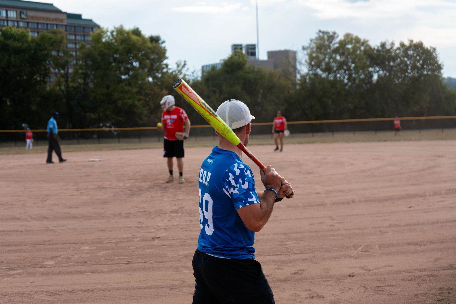 A softball player in a blue jersey winds up to bat on a dirt field during an outdoor game.