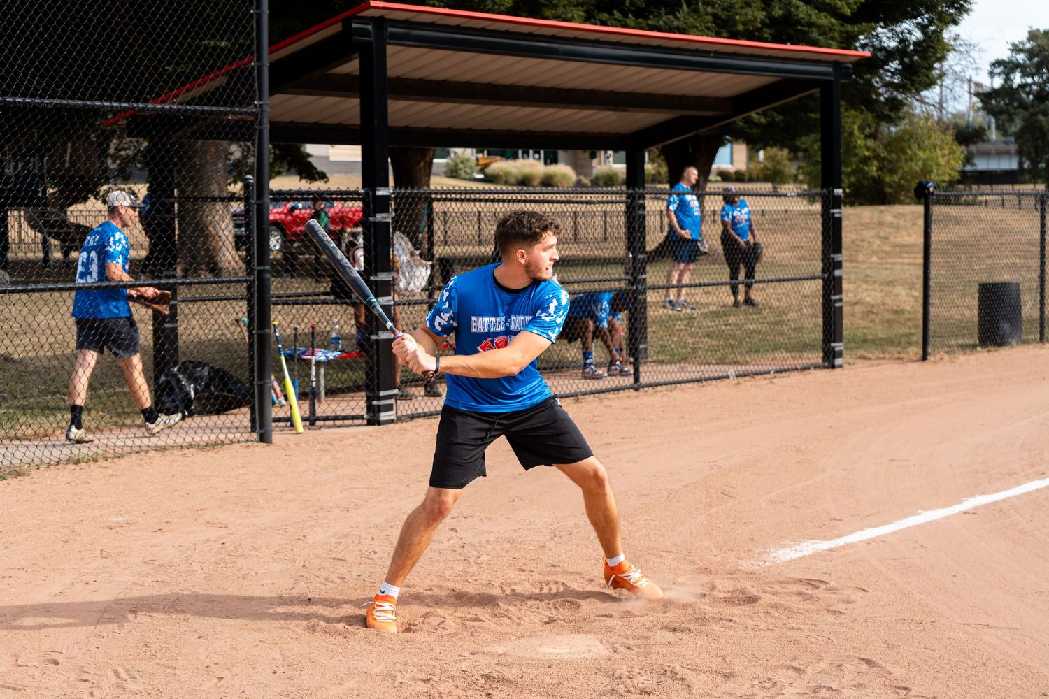A male softball player swings a bat at home plate during an outdoor game on a dirt field.